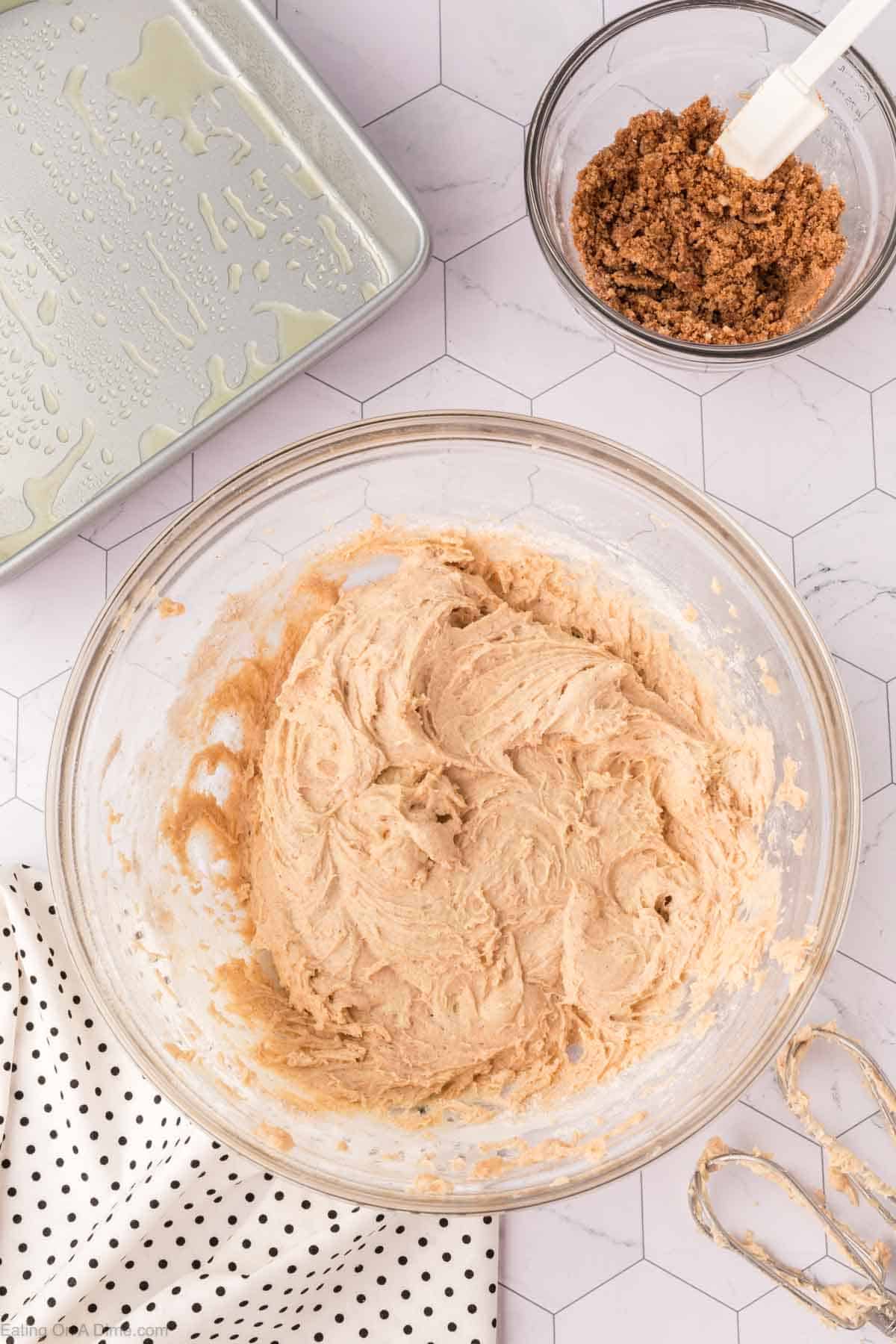 A glass bowl filled with creamy brown batter for a delicious coffee cake recipe sits on a hexagonal patterned surface. Nearby is a bowl of crumbly brown sugar topping, a greased baking pan, and a polka dot cloth. An electric mixer beater is partially visible.