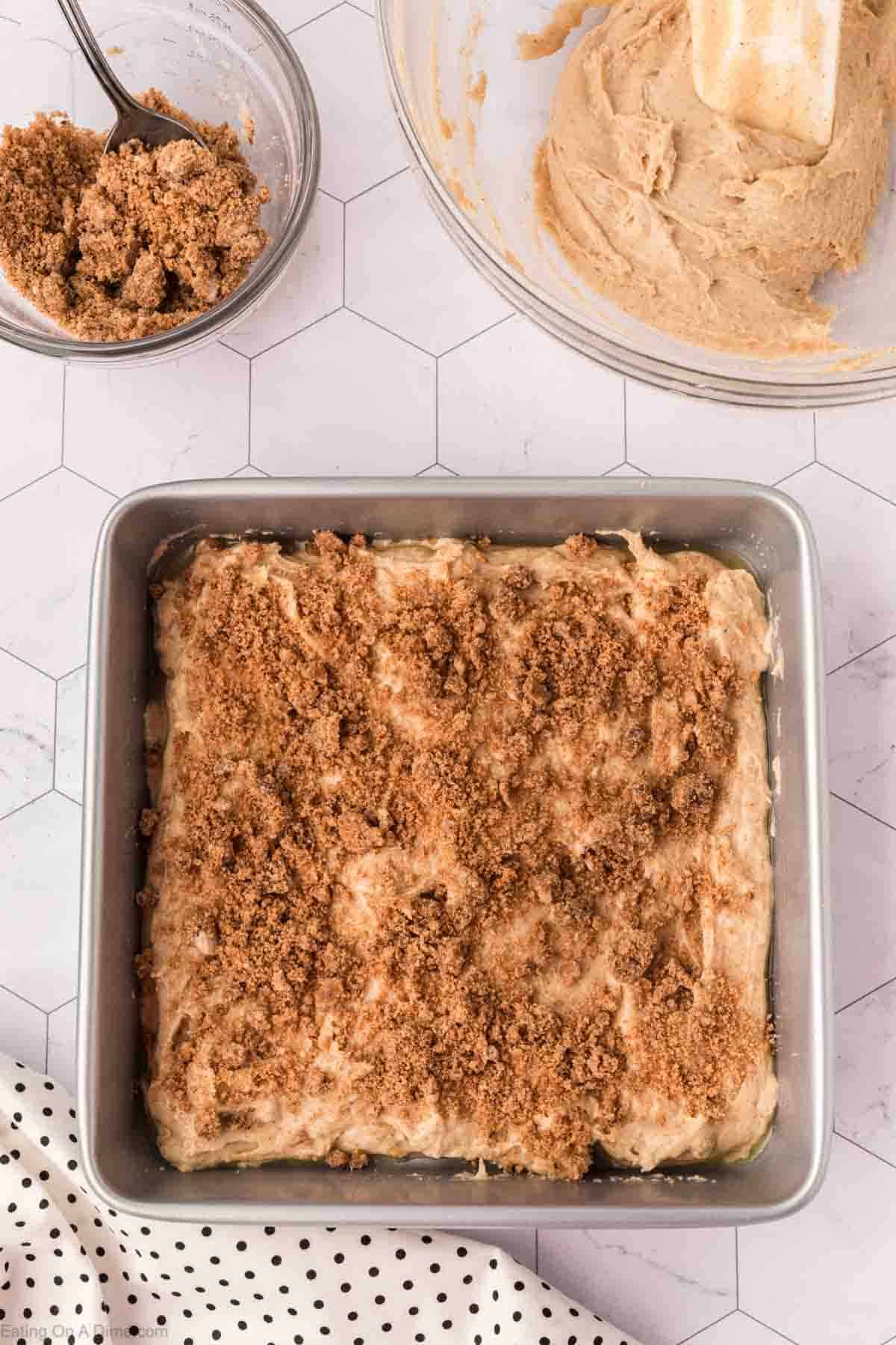 A square baking pan brims with cinnamon coffee cake mixture, crowned by a brown crumbly streusel. Nearby sits a small bowl of extra brown sugar streusel and a large bowl holding more batter from this delightful coffee cake recipe. The surface features a white hexagonal pattern.