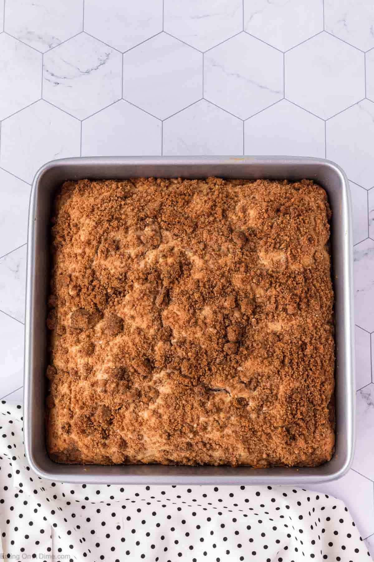 A square coffee cake with a crumbly cinnamon topping sits invitingly in a metal baking pan, hinting at the delicious coffee cake recipe within. The pan is on a hexagonal-patterned surface, with a white and black polka dot cloth peeking from the bottom left corner.