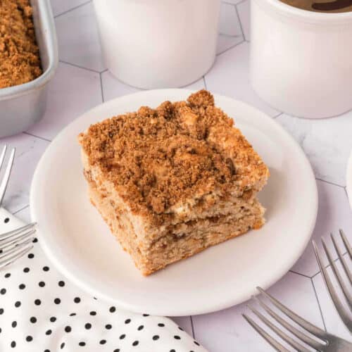 A slice of crumb cake, reminiscent of a classic coffee cake recipe, rests on a white plate against a hexagonal tile surface. The cake features a golden brown crumbly layer. Nearby, you'll find a white napkin with black dots, several forks, and white containers with lids.