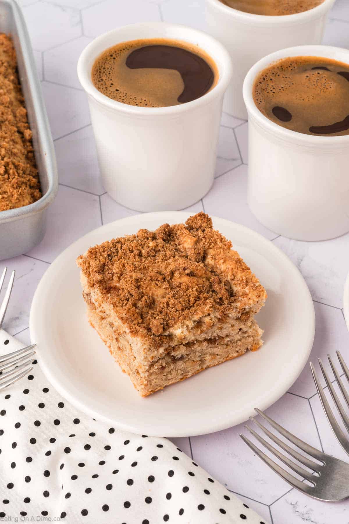 A slice of crumb coffee cake, inspired by a beloved coffee cake recipe, rests on a white plate amid cups of coffee and a polka dot napkin. Nearby, silver forks await use while a baking tray with more delicious cake beckons in the background.