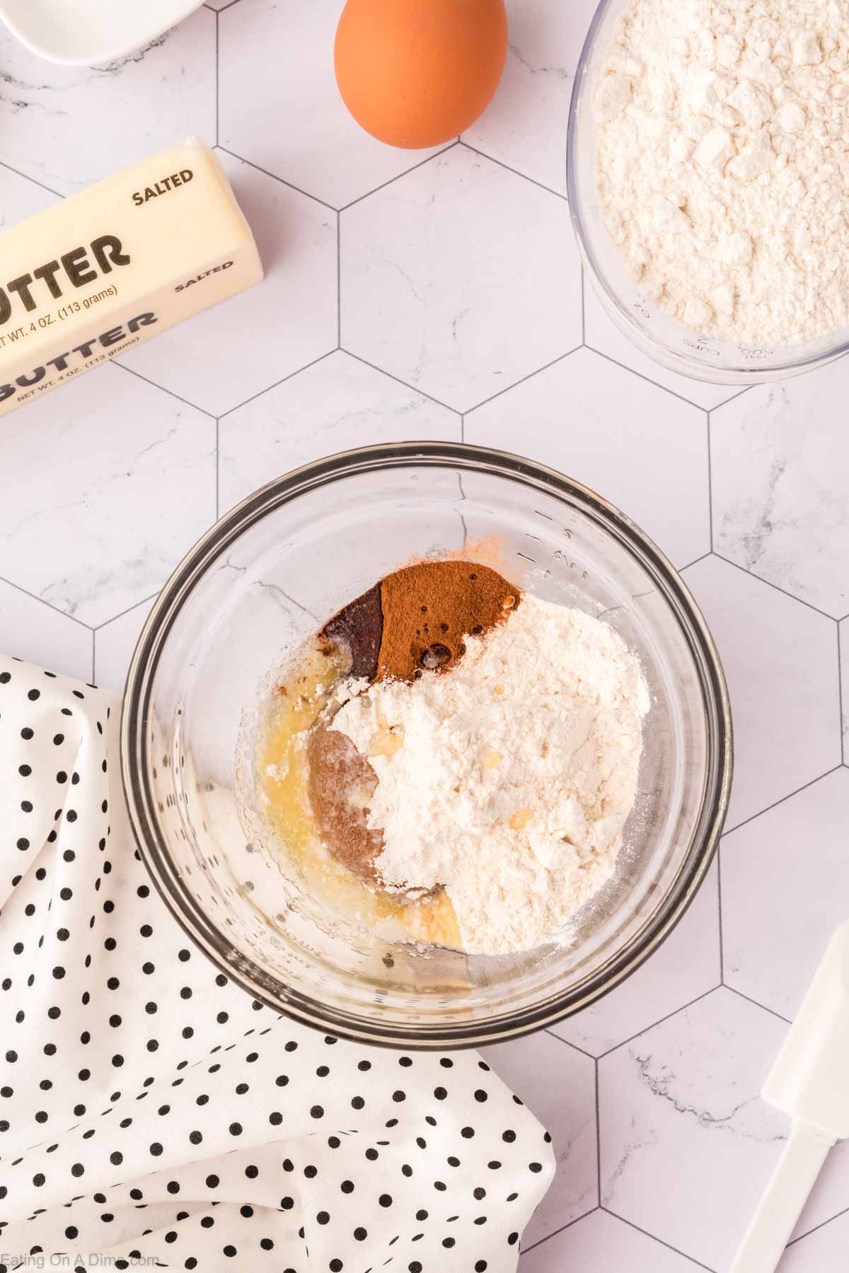 A glass mixing bowl holds flour, melted butter, cocoa powder, and sugar, poised for blending into a delicious treat. Nearby are a stick of salted butter, an egg, and a polka dot kitchen towel on a hexagonal-tiled surface—essentials for crafting the perfect coffee cake recipe.