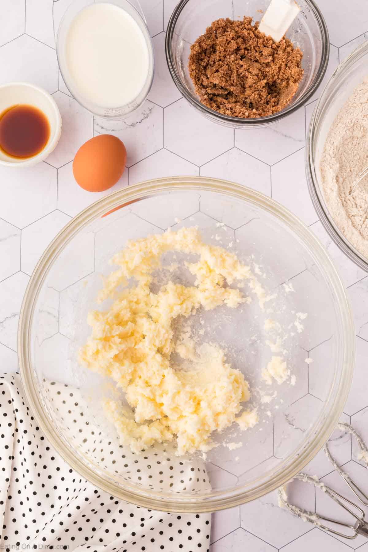 Ingredients for baking a coffee cake are arranged on a hexagonal patterned surface. A clear bowl holds creamed butter and sugar. Nearby are bowls of brown sugar, flour, and vanilla, alongside an egg and a cup of milk. A polka dot cloth and whisk rest beside them, ready for the recipe adventure.