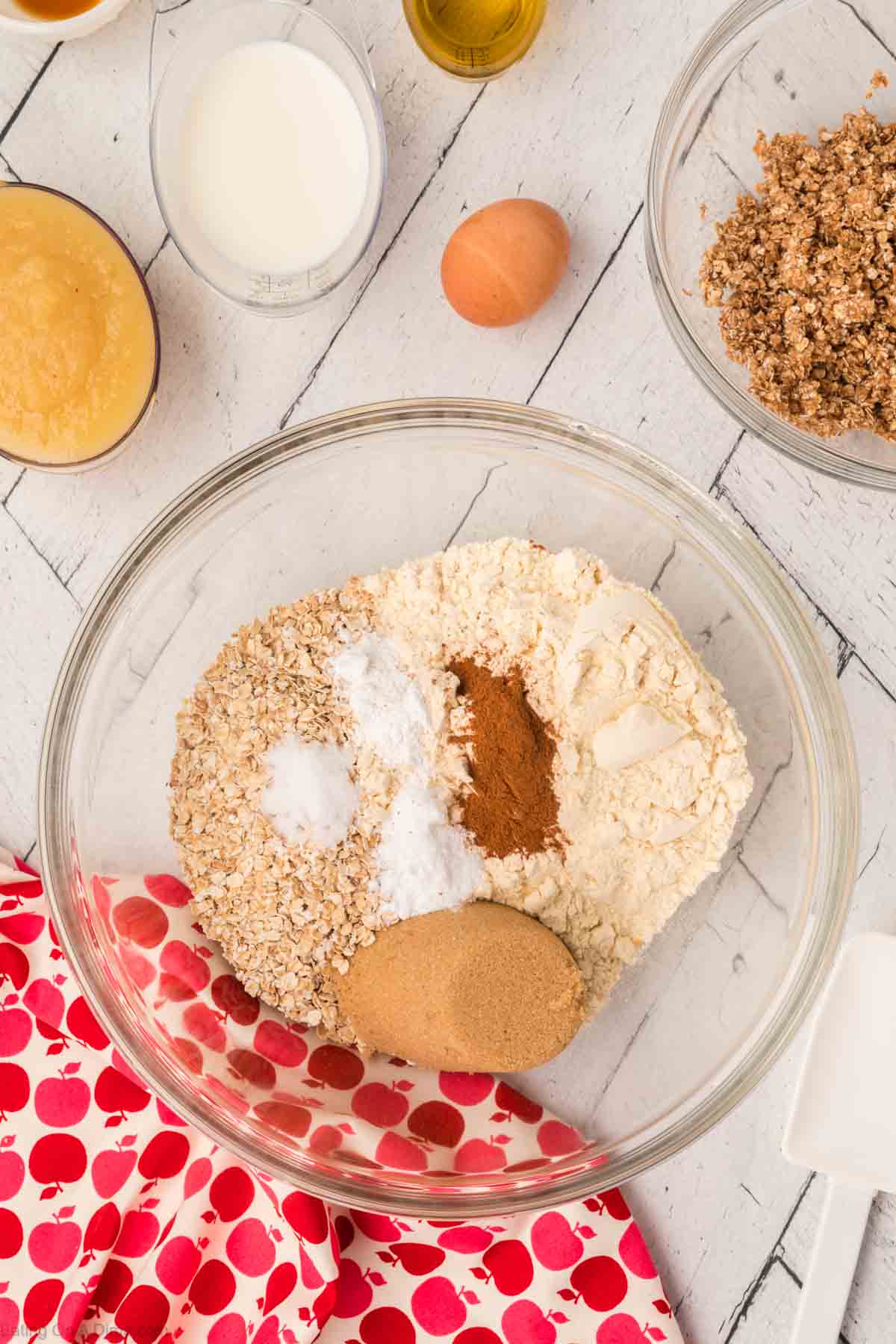 Baking ingredients on a countertop include rolled oats, flour, brown sugar, cinnamon, and baking soda in a clear bowl—all part of an oatmeal applesauce muffins recipe. Nearby are bowls of applesauce, milk, and a crumb mixture. An egg and a red and white polka-dot cloth are also visible.