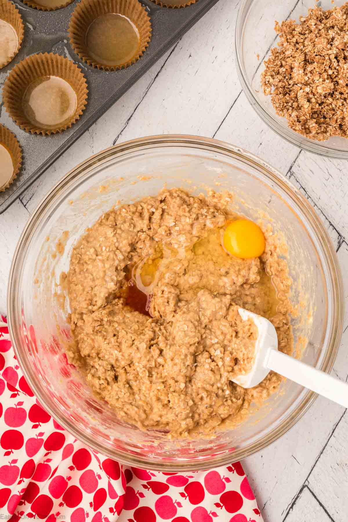 A glass mixing bowl filled with batter mixture, a raw egg, and a white spatula on a white wooden surface. Beside it is a red and white polka dot cloth, a bowl with crumbly mixture for an oatmeal applesauce muffins recipe, and a muffin tray lined with brown paper muffin cups.