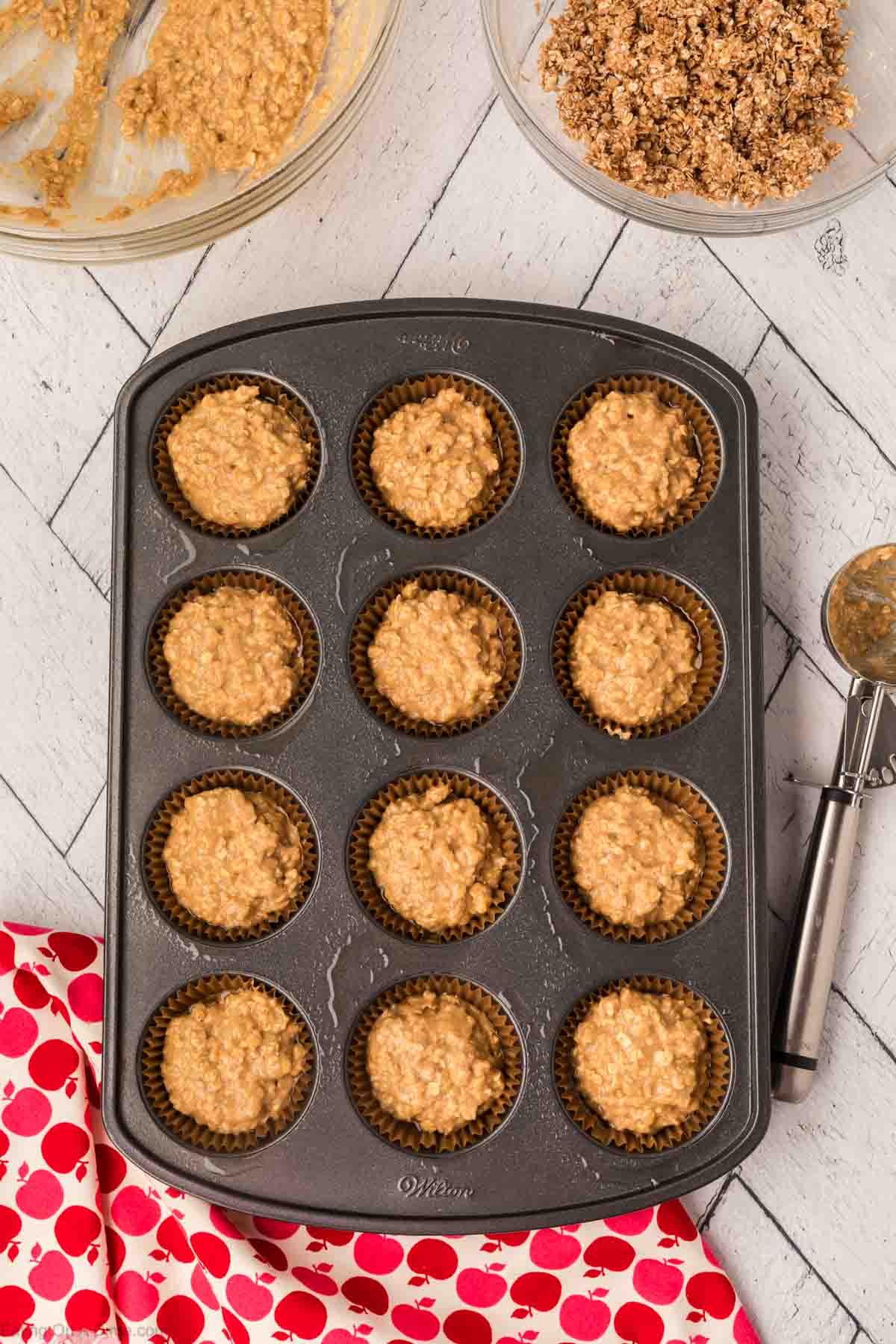 A muffin tray filled with unbaked oatmeal applesauce muffins batter sits on a white wooden surface. Next to it is a large bowl with leftover batter, a smaller bowl with crumble topping, and a metal scoop. A red cloth with white and pink cherries is partially visible on the side.