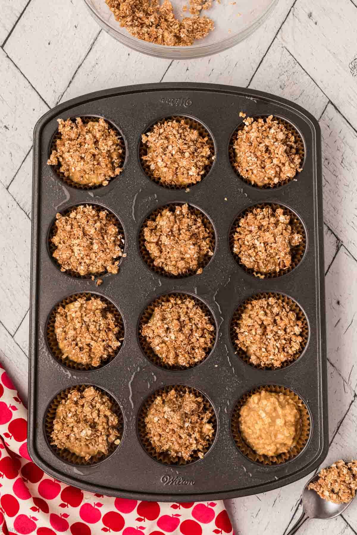 A muffin tin filled with 12 unbaked oatmeal applesauce muffins, some with a crumbly topping, is placed on a light-colored, patterned surface. A red cloth with white polka dots is partially visible in the bottom left corner, and a metal spoon is on the right.