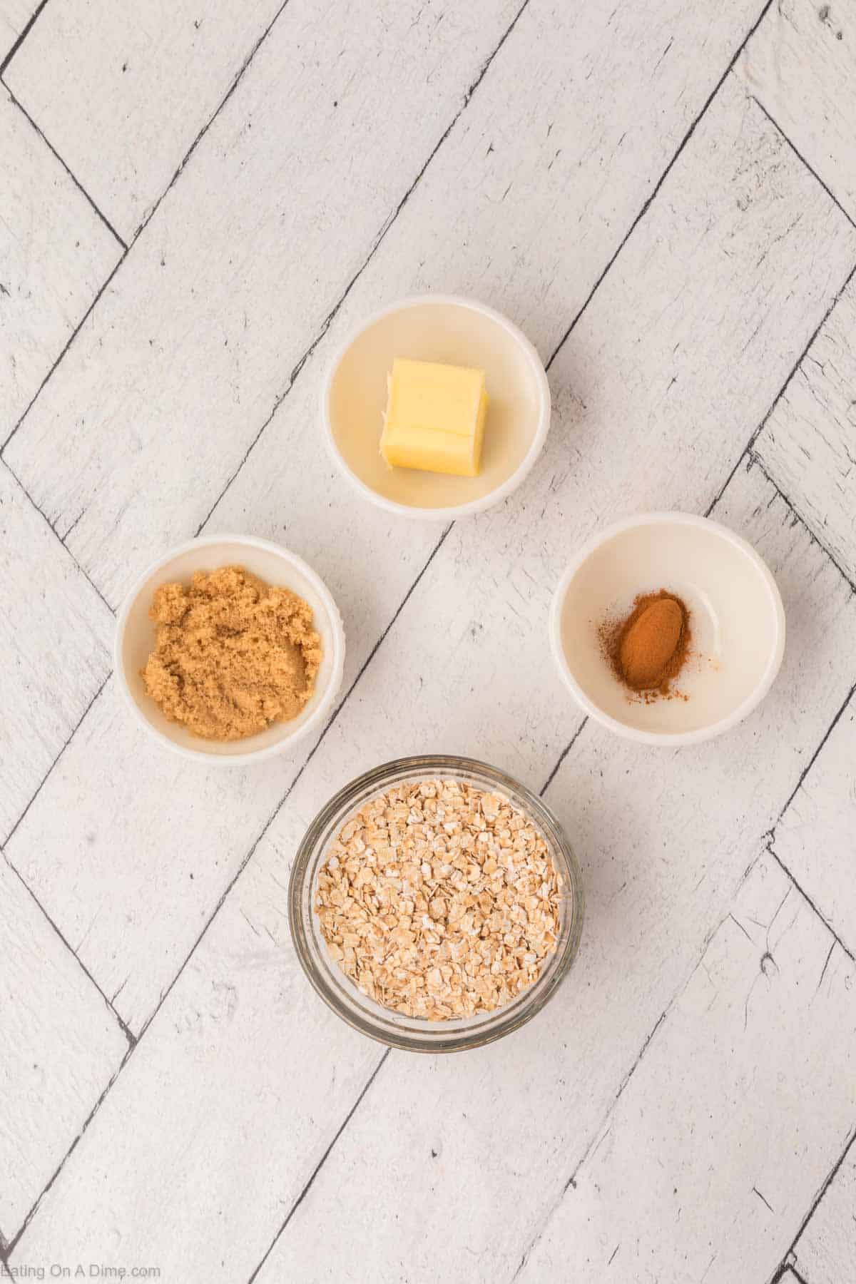 A flat lay photo on a white wooden surface showing four small dishes containing ingredients. There is a small glass jar of rolled oats, a bowl of brown sugar, a bowl with a pat of butter, and a bowl of ground cinnamon—perfect for crafting an oatmeal applesauce muffins recipe.