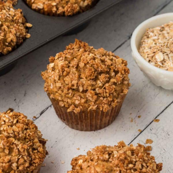 A close-up of an oatmeal applesauce muffin topped with a crumbly oat streusel sits on a rustic wooden surface. Other muffins in a muffin tin and a small bowl of oats are visible in the background, highlighting the cozy, homemade pastry setting suggested by this delightful recipe.