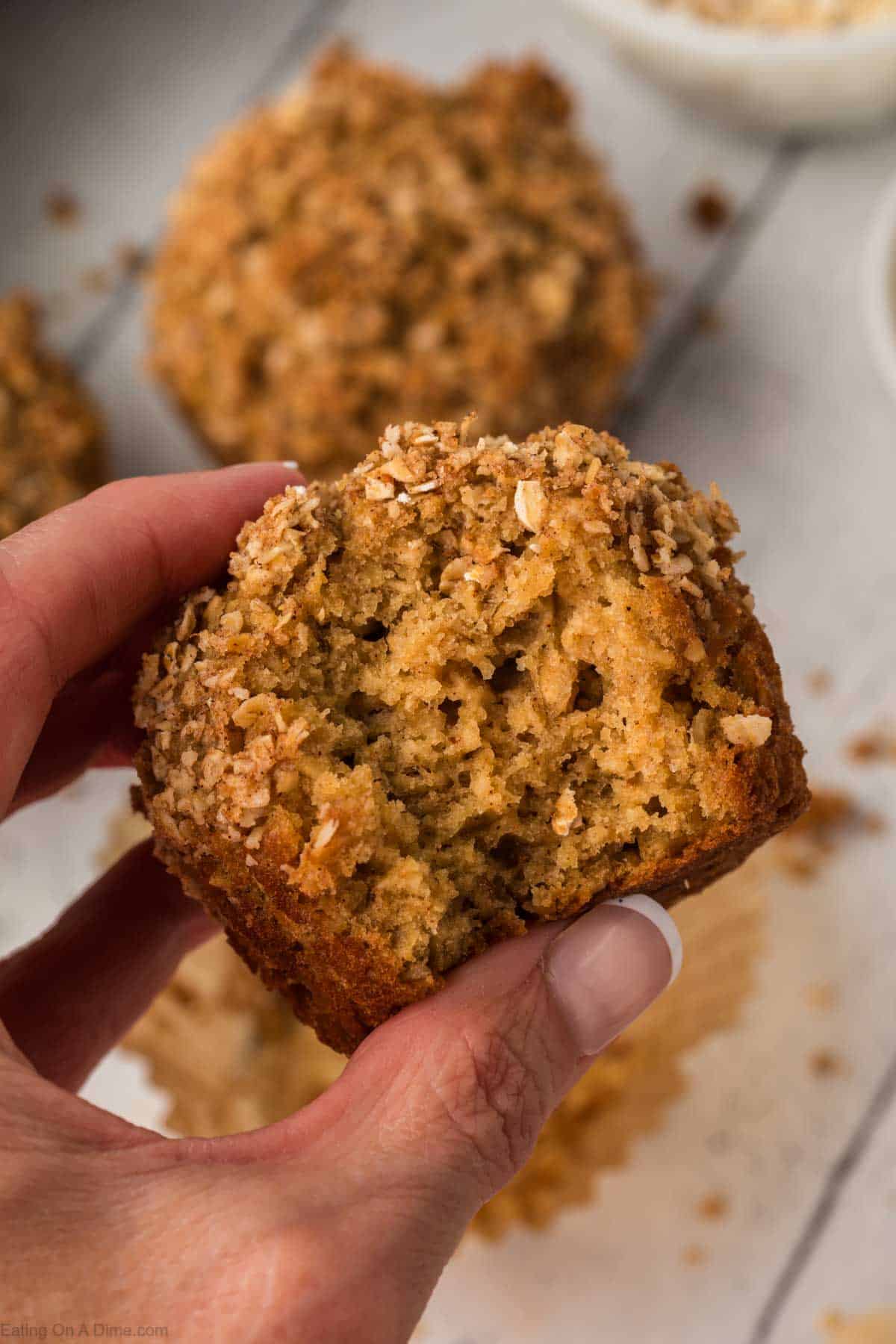 A hand holding a crumbly, golden-brown oatmeal applesauce muffin topped with oats and crumbs, with more muffins blurred in the background. The muffin appears moist and freshly baked.