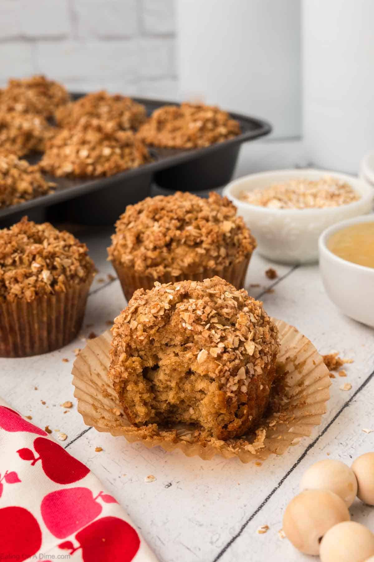 A close-up of an oatmeal applesauce muffin with a bite taken out, revealing its moist interior. The muffin is placed in a partially opened paper liner on a white wooden surface. Other muffins in a muffin tin, bowls of oats, and a red and white cloth are in the background, perfect for any recipe.