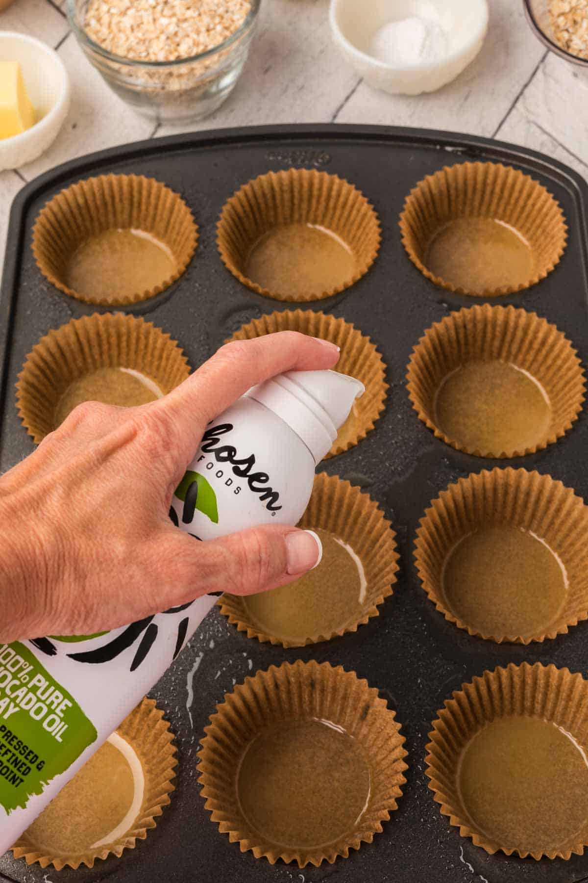 A hand is holding and spraying a can of avocado oil into a lined muffin pan for an oatmeal applesauce muffins recipe. The muffin cups are made of brown paper, and eight out of twelve are empty. The background shows other baking ingredients such as oats, sugar, and butter.