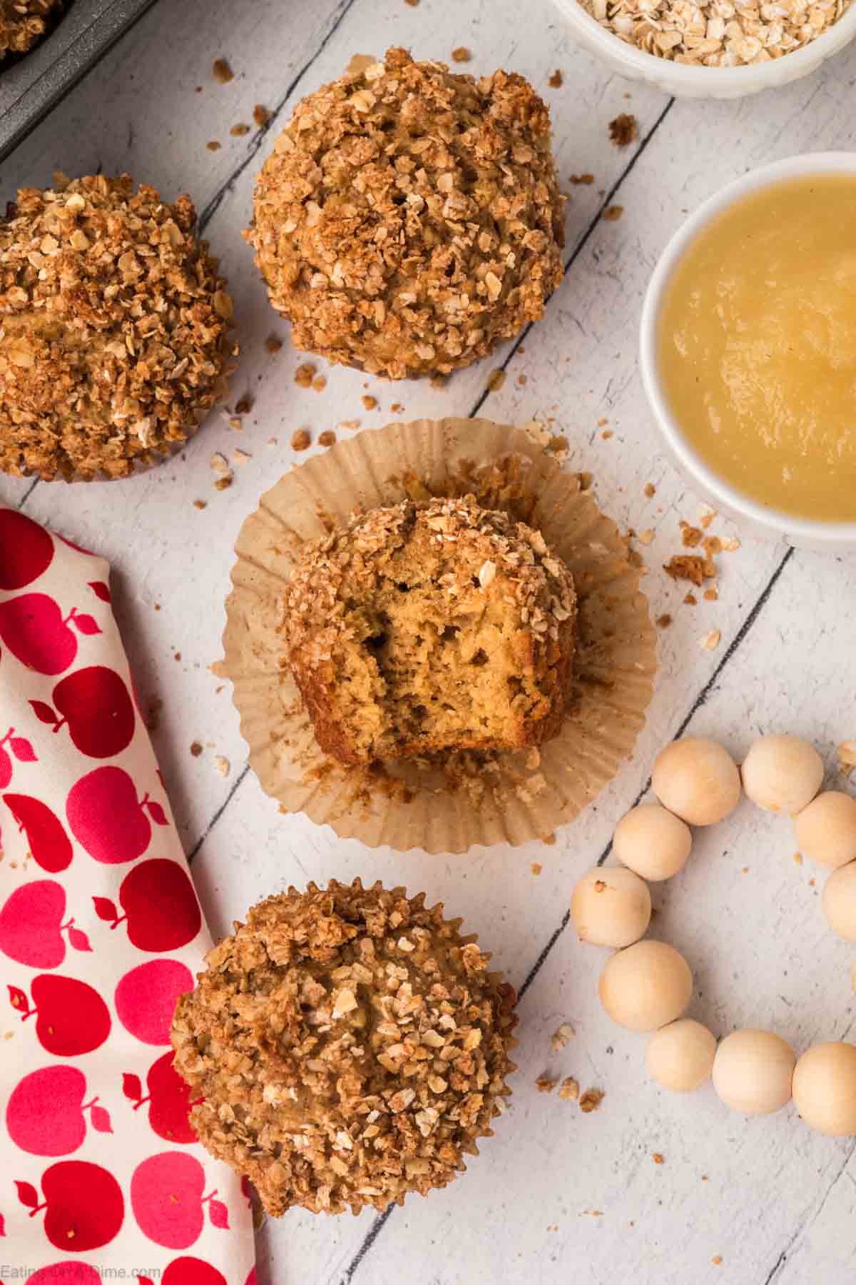 Top view of freshly baked, crumbly oatmeal applesauce muffins surrounded by oats on a white wooden surface. One muffin is half unwrapped, revealing its moist, fluffy texture. A small bowl of applesauce, a red and white apple-patterned cloth, and wooden beads are nearby.