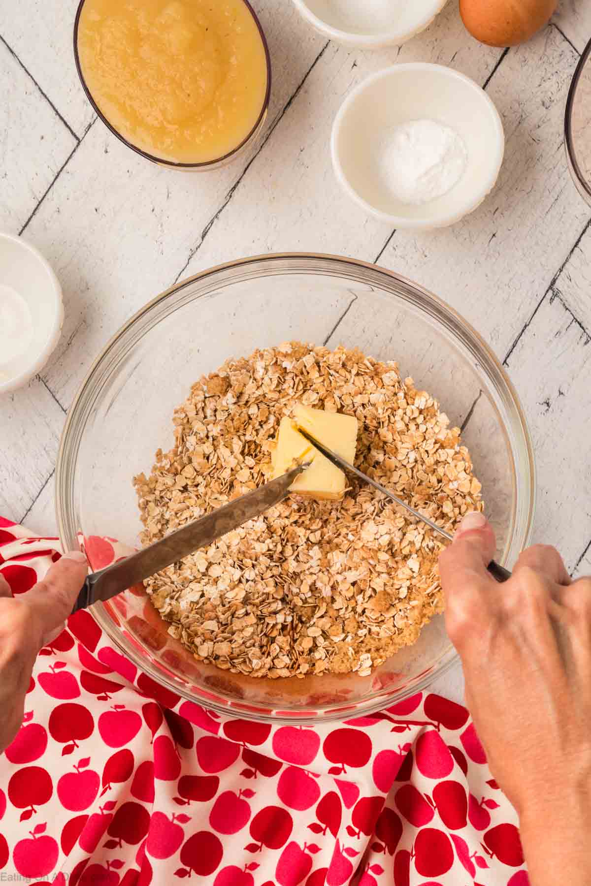 Two hands use knives to cut a block of butter into a glass bowl filled with oats, prepping for an oatmeal muffins recipe. Surrounding the bowl are small dishes containing various ingredients. A red and white patterned cloth is partially visible in the bottom left corner on a white wooden surface.