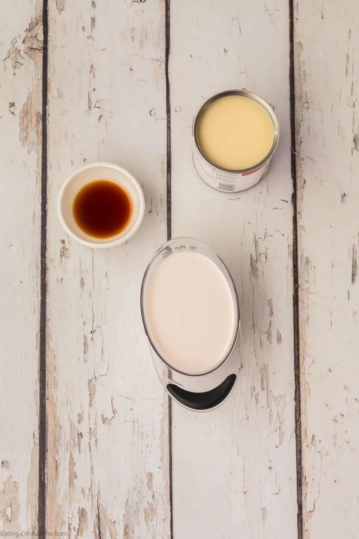 A top-down view of three ingredients on a white rustic wooden surface, perfect for homemade coffee creamer. There is a small white bowl containing vanilla extract, an open can of sweetened condensed milk, and a measuring cup filled with cream.