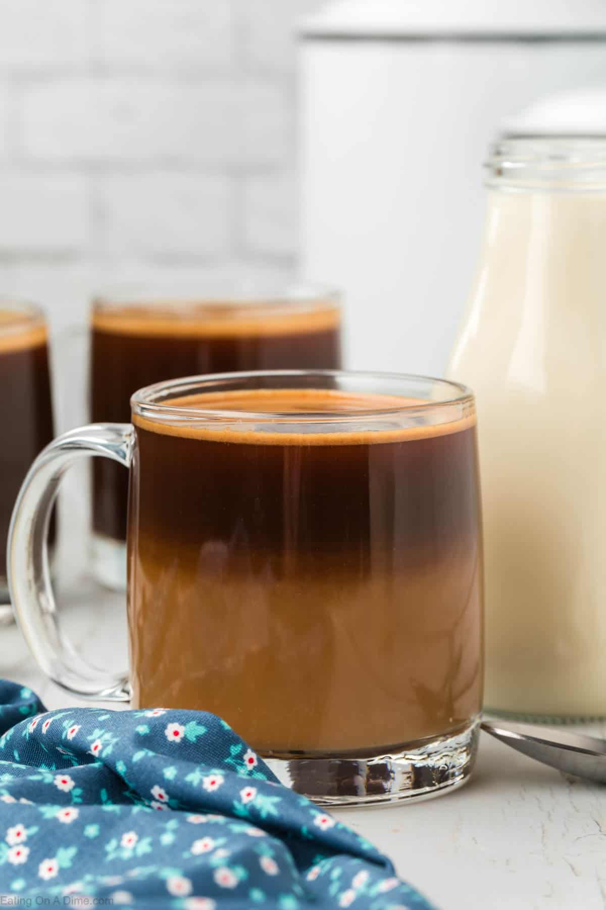 A clear glass mug is filled with layered iced coffee, showcasing dark coffee on top and light cream at the bottom. Another mug of layered coffee and a glass bottle of homemade coffee creamer are in the background. A blue floral cloth is partially draped beside the mug.