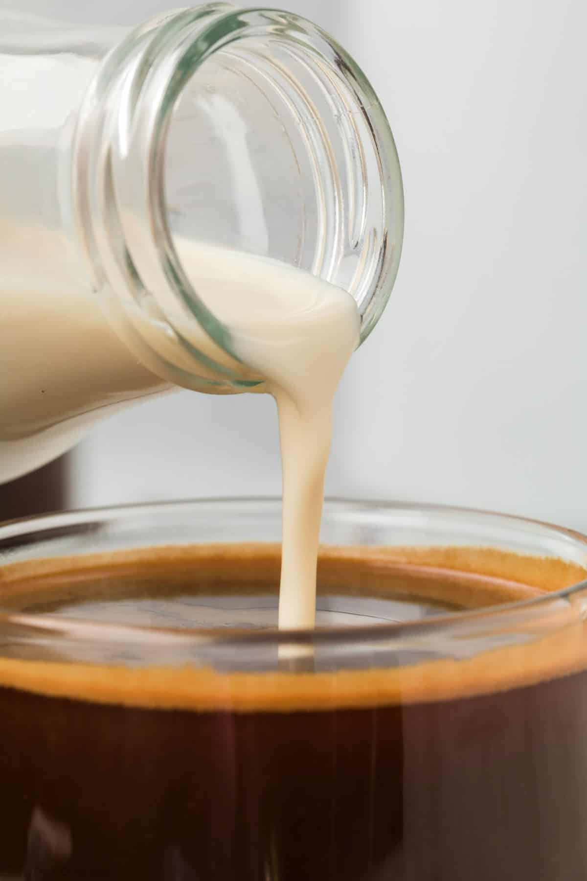 Close-up of homemade coffee creamer being poured from a glass bottle into a cup containing dark coffee. The creamy stream mingles with the rich coffee, creating a striking contrast between the light cream and the dark brew. The background is blurred and neutral-toned.