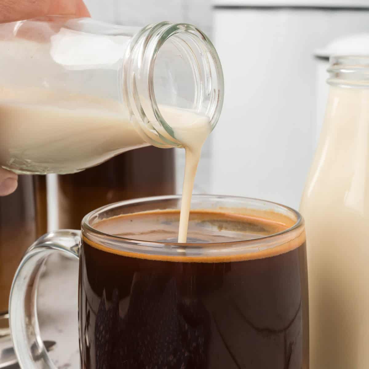 Close-up of a hand pouring milk from a glass bottle into a mug of black coffee. Another bottle of milk is visible in the background. The scene suggests adding homemade coffee creamer to enhance your perfect cup.