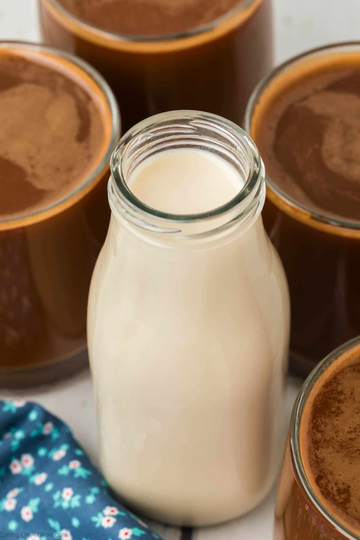 A glass bottle filled with milk, resembling a homemade coffee creamer, is surrounded by several mugs of coffee. The bottle is centrally positioned, contrasting against the dark liquid in the mugs. A blue cloth with a floral pattern is partially visible at the bottom left corner.