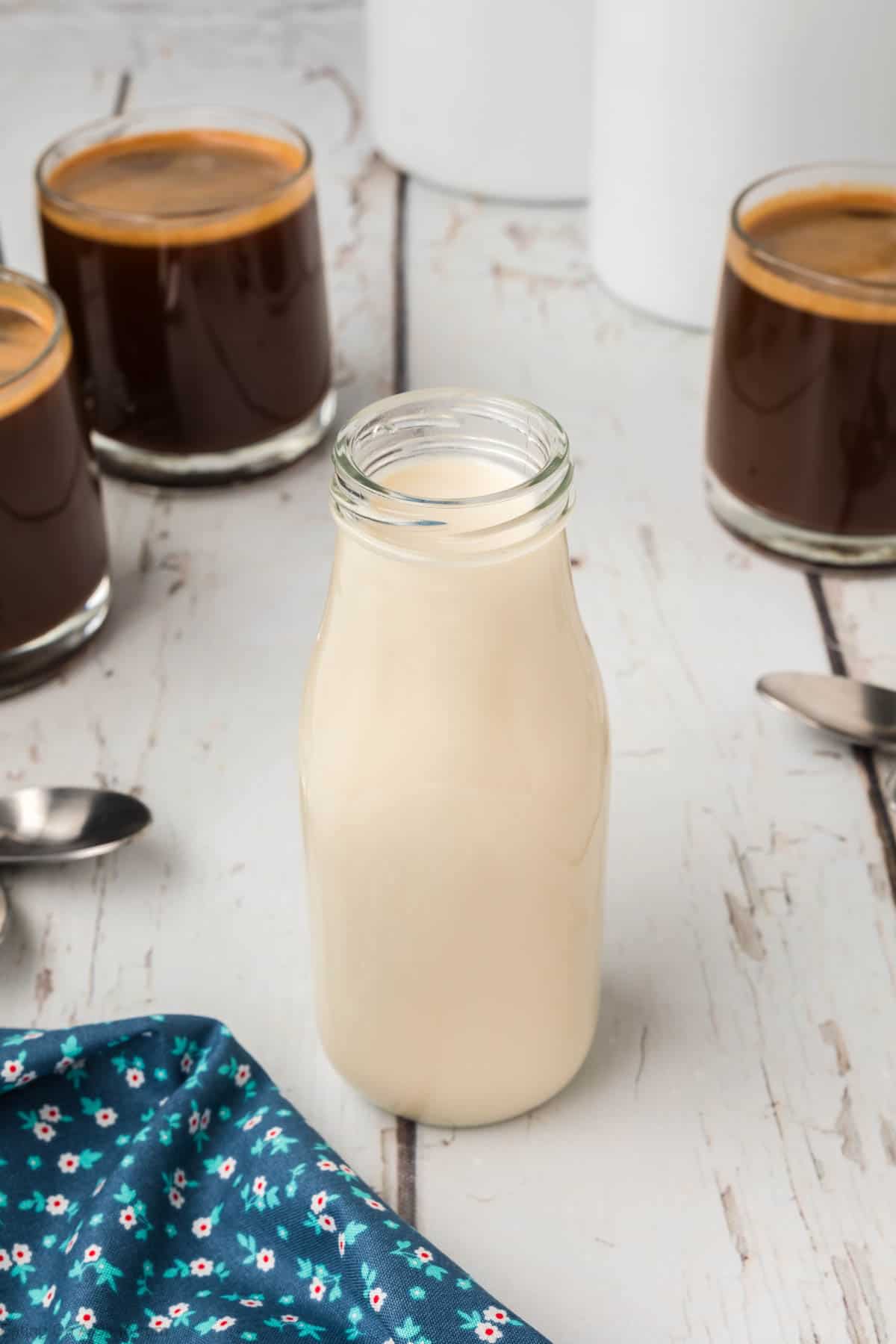 A glass bottle of milk is placed on a wooden table. Surrounding it are three small glasses filled with dark coffee and homemade coffee creamer. Two spoons lie on the table, and a blue floral cloth is partially visible in the bottom left corner.