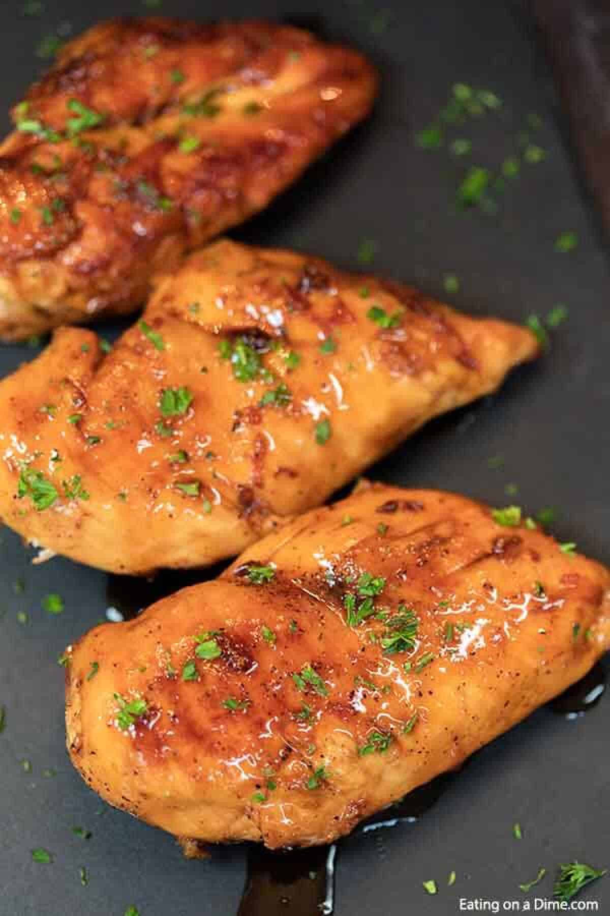 Three grilled chicken breasts, expertly glazed with a shiny brown sugar sauce, are garnished with chopped herbs and beautifully presented on a dark platter.