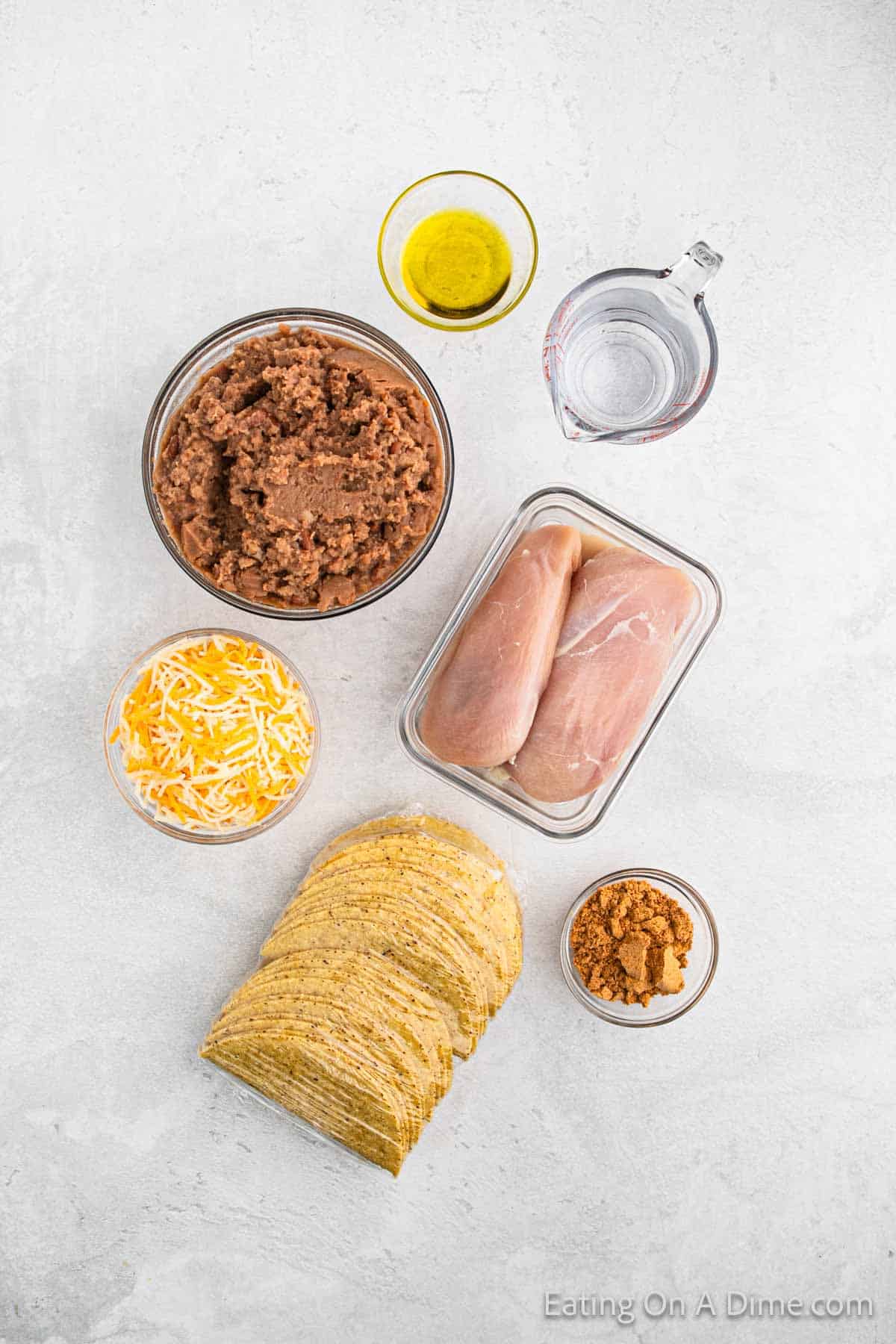 Overhead view of taco ingredients on a white surface: a bowl of refried beans, raw chicken breasts in a dish ready for baked chicken tacos, shredded cheese, stacked taco shells, seasonings, and cups of water and oil.