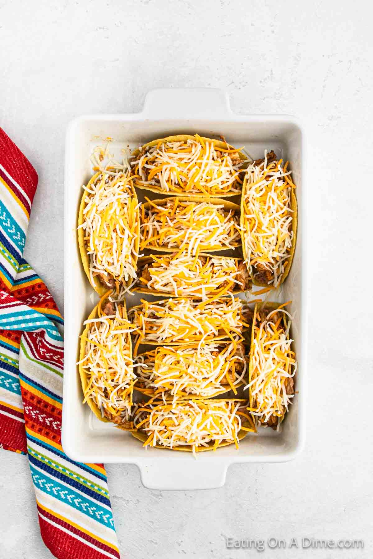 A white baking dish showcases six hard shell tacos filled with shredded beef and baked to perfection. Topped with a generous amount of melted cheese, they rest beside a colorful, striped fabric on a light surface. These delightful treats evoke the flavors of classic baked chicken tacos.