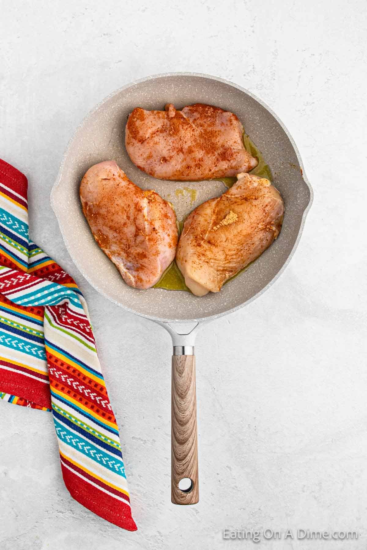 A pan with three seasoned chicken breasts cooking in oil sits on a light countertop, reminiscent of sizzling ingredients for baked chicken tacos. Next to the pan is a colorful, striped kitchen towel.