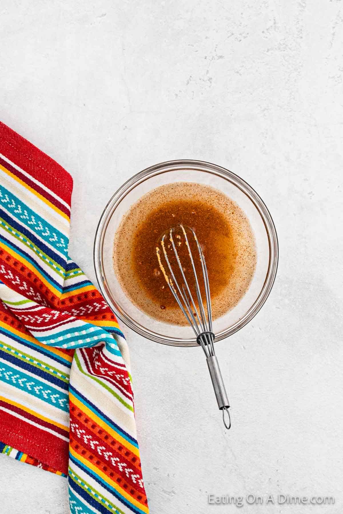 A clear glass bowl with a brown liquid mixture and a metal whisk rests on a textured white surface, ready to marinate the Baked Chicken Tacos. To the side, a colorful striped kitchen towel with red, blue, green, yellow, and white patterns is partially visible.