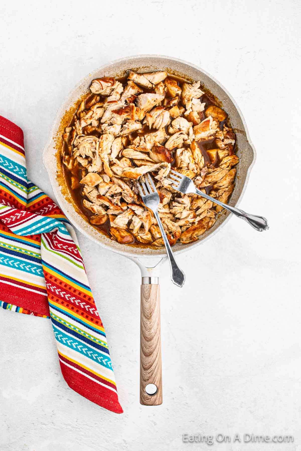 A skillet with shredded chicken in sauce, perfect for baked chicken tacos, accompanied by two forks. A colorful striped cloth is next to the pan.