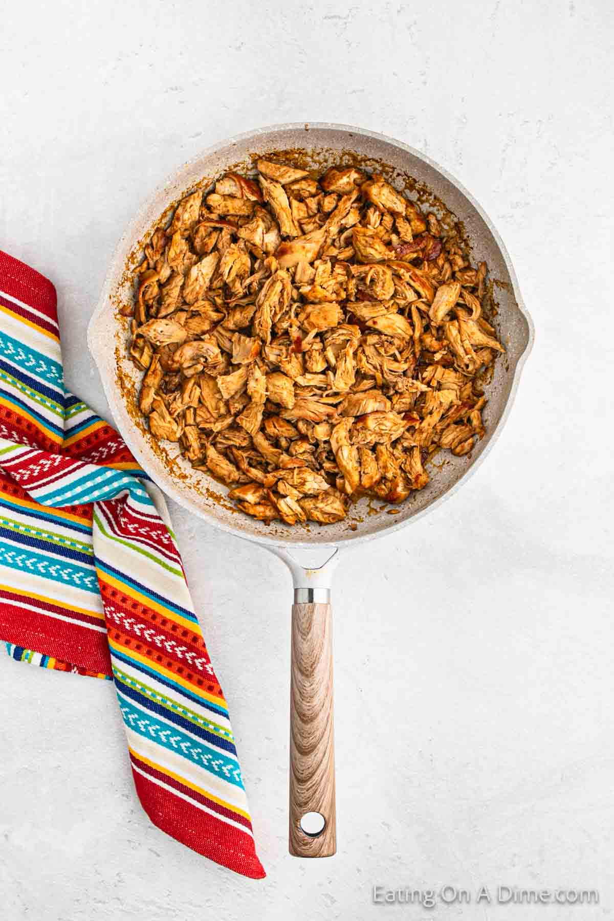 A frying pan filled with seasoned, shredded chicken, perfect for Baked Chicken Tacos. A colorful, striped cloth is placed next to the pan on a light gray surface.