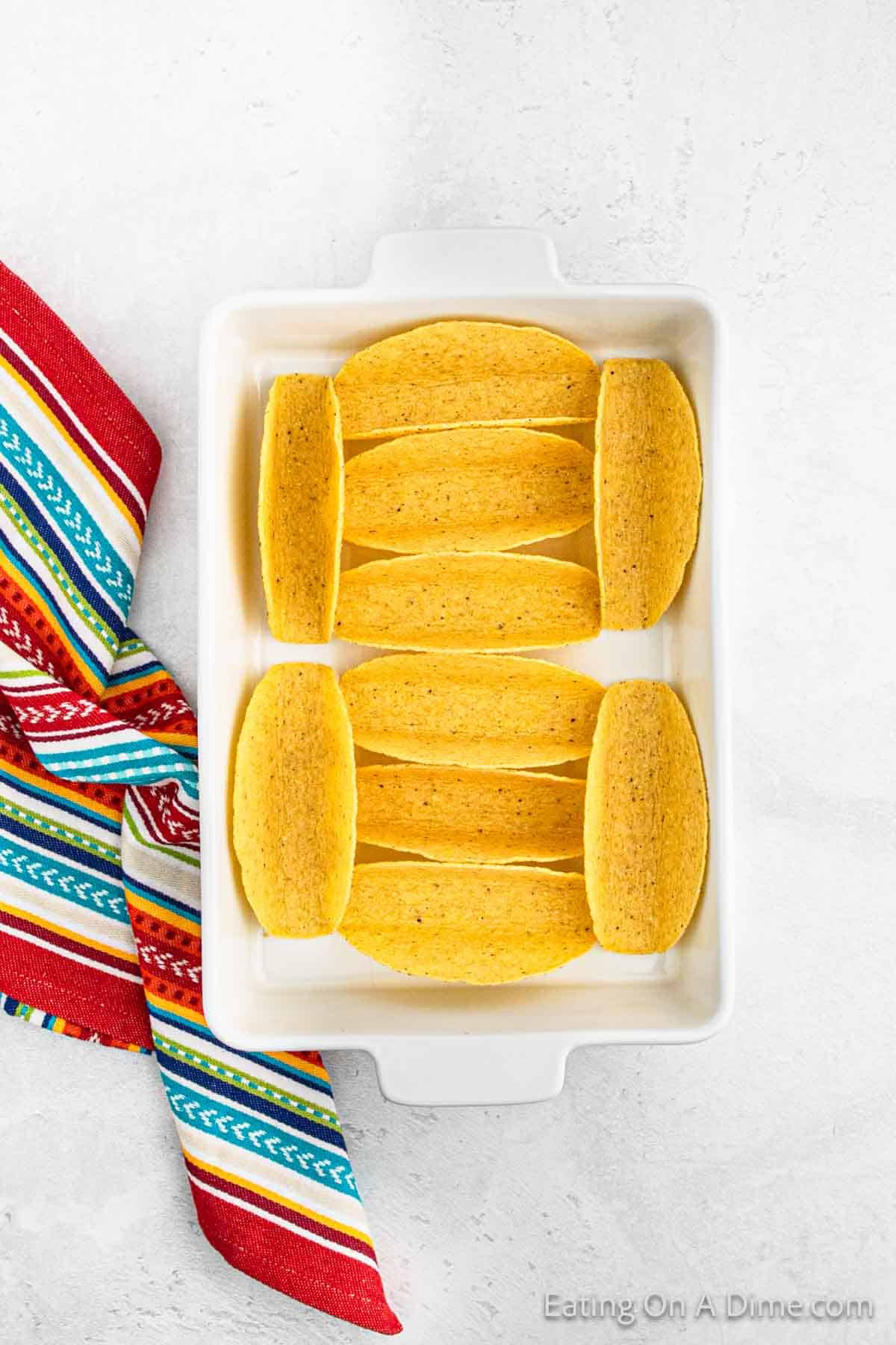 Empty yellow taco shells are neatly arranged in a white baking dish, ready for delicious Baked Chicken Tacos. A colorful striped cloth with red, blue, and green patterns is partially visible on the side. The dish rests elegantly on a light gray surface.