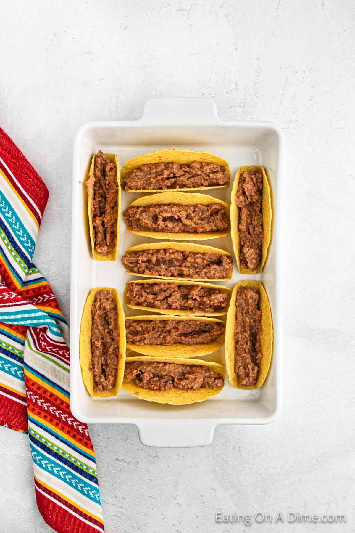 A white rectangular baking dish filled with hard taco shells stuffed with seasoned ground beef, evoking the flavor of baked chicken tacos. The dish rests beside a colorful striped cloth napkin on a light gray surface.