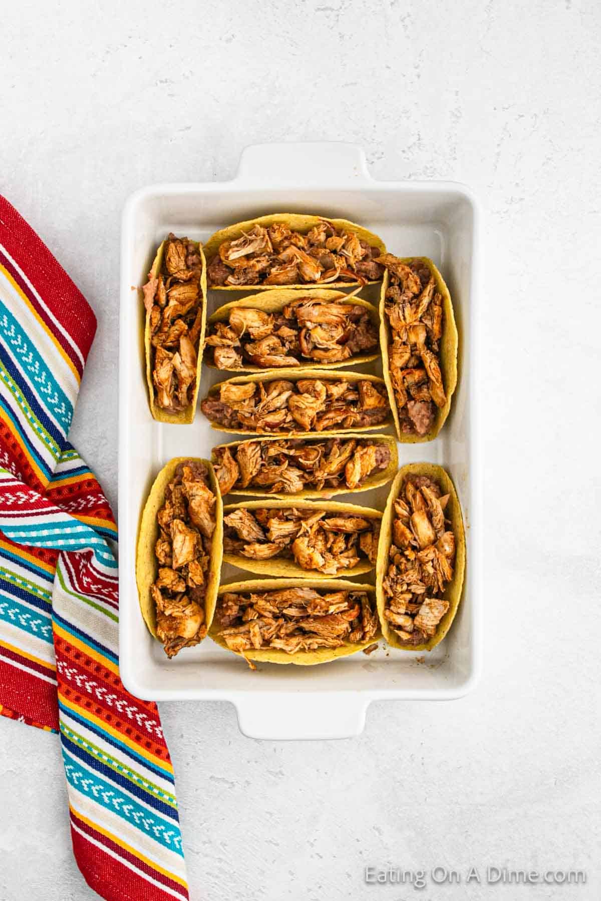 A white baking dish brimming with eight hard-shell baked chicken tacos, each generously stuffed with shredded chicken. The vibrant meal is set on a textured white surface, perfectly complemented by a colorful striped cloth adorning the side.