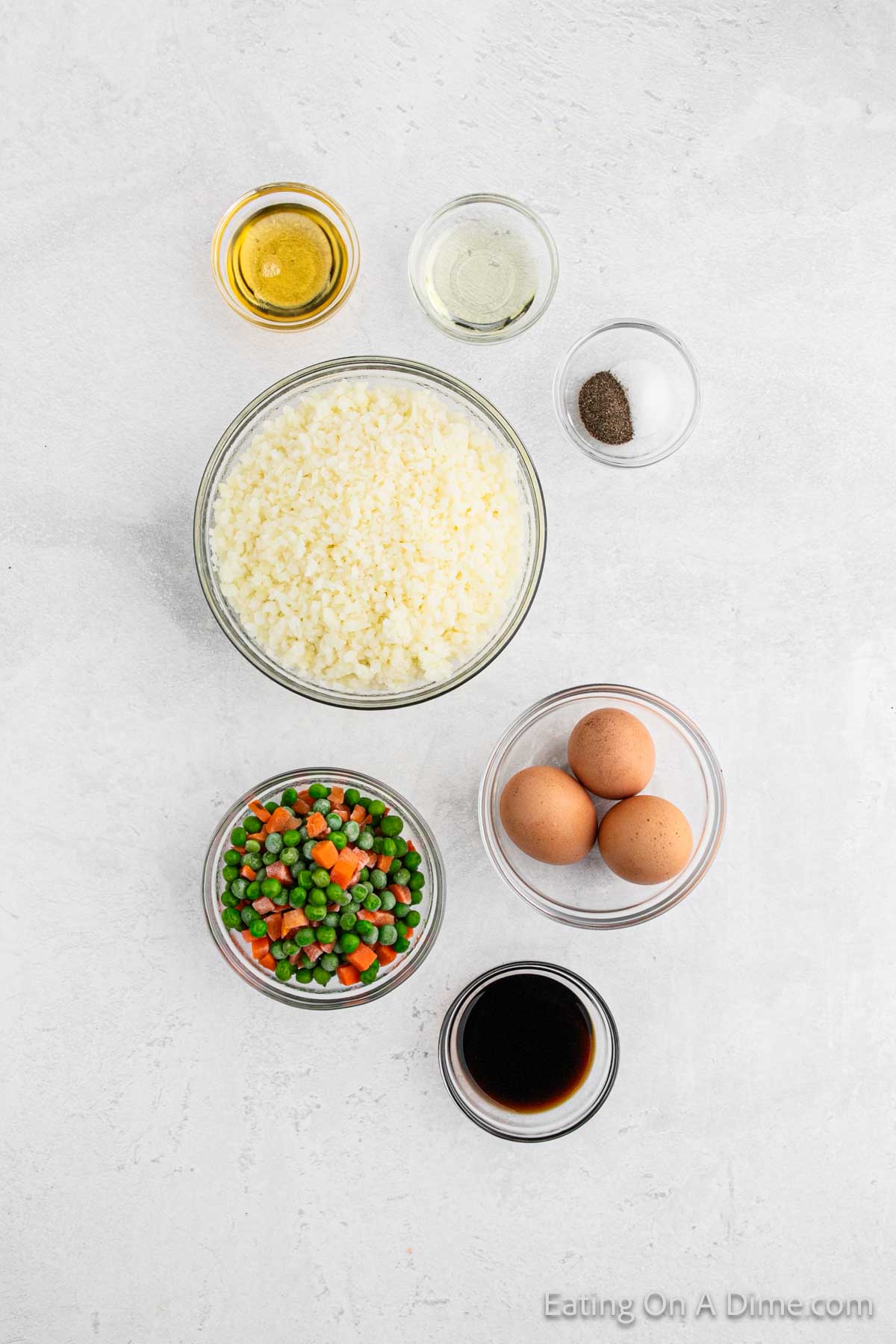 Ingredients for a delightful cauliflower fried rice arranged on a white surface: a bowl of cauliflower rice, mixed peas and carrots, three eggs, small bowls of oil, vinegar, soy sauce, and a bowl with salt and pepper.