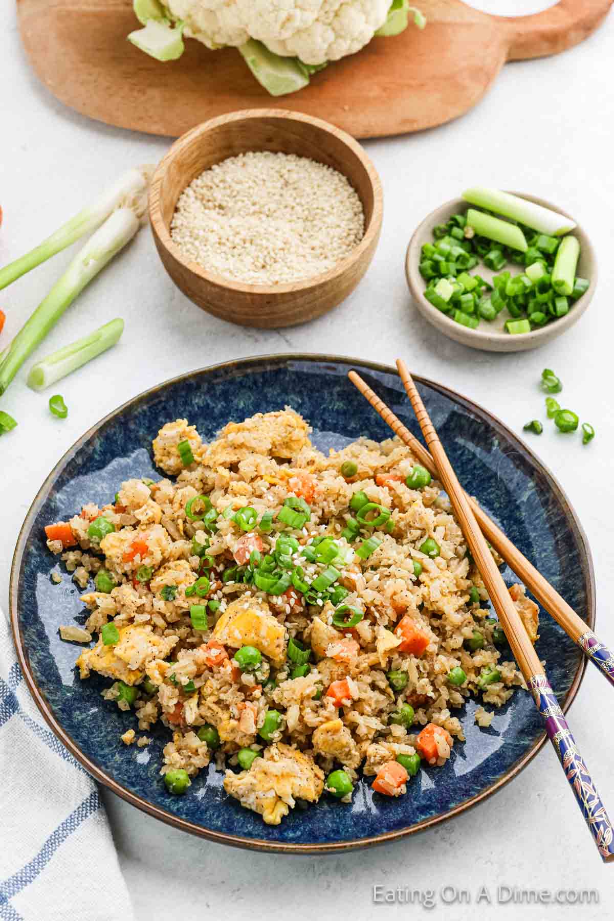 A blue plate showcases cauliflower fried rice with peas, carrots, and scrambled eggs, garnished with chopped green onions. Chopsticks rest on the plate. Nearby are bowls of sesame seeds and more green onions on a white surface, completing this delicious cauliflower fried rice dish.