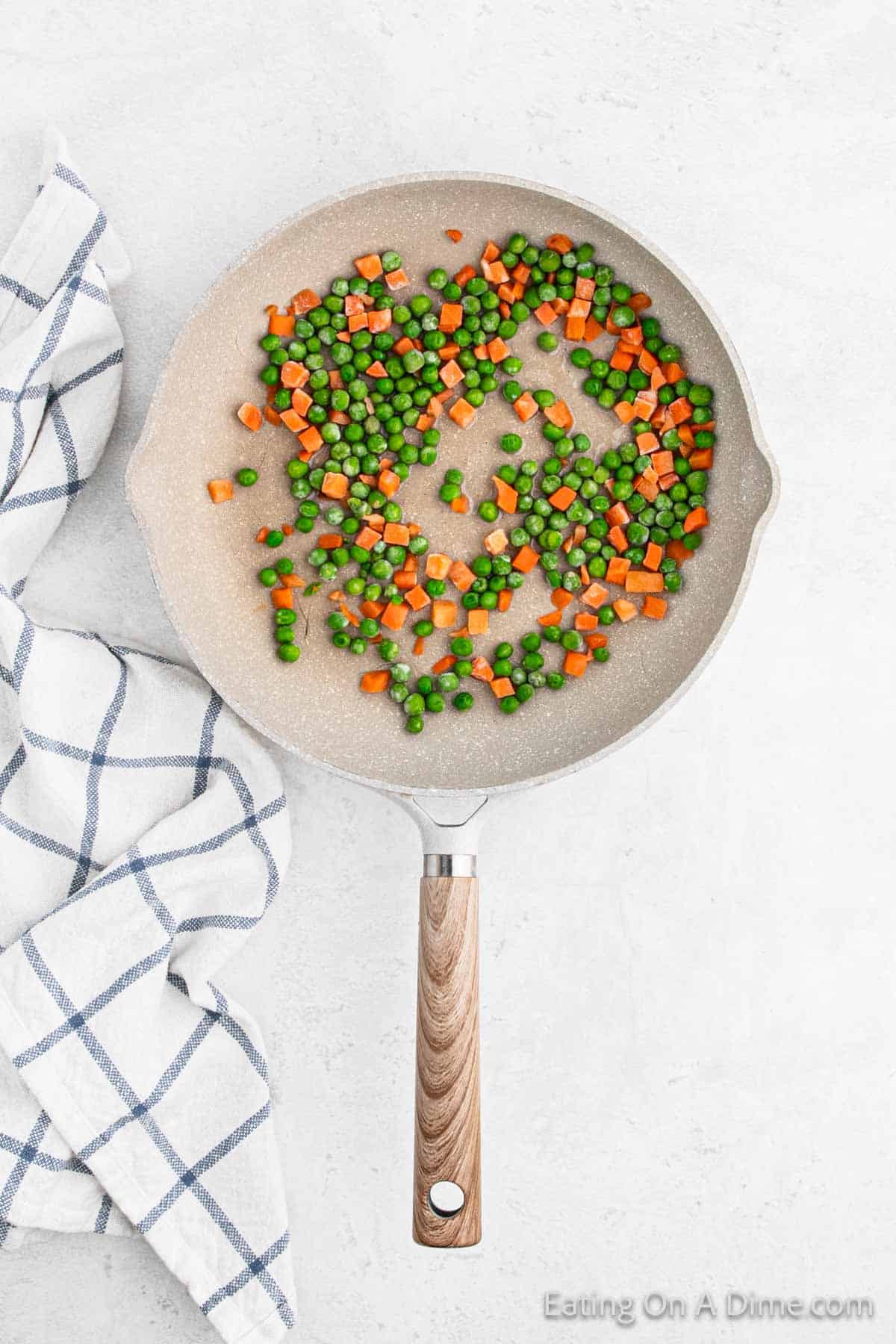 A frying pan with mixed peas and diced carrots sits on a light surface next to a white and blue checkered cloth, featuring a delicious serving of cauliflower fried rice.