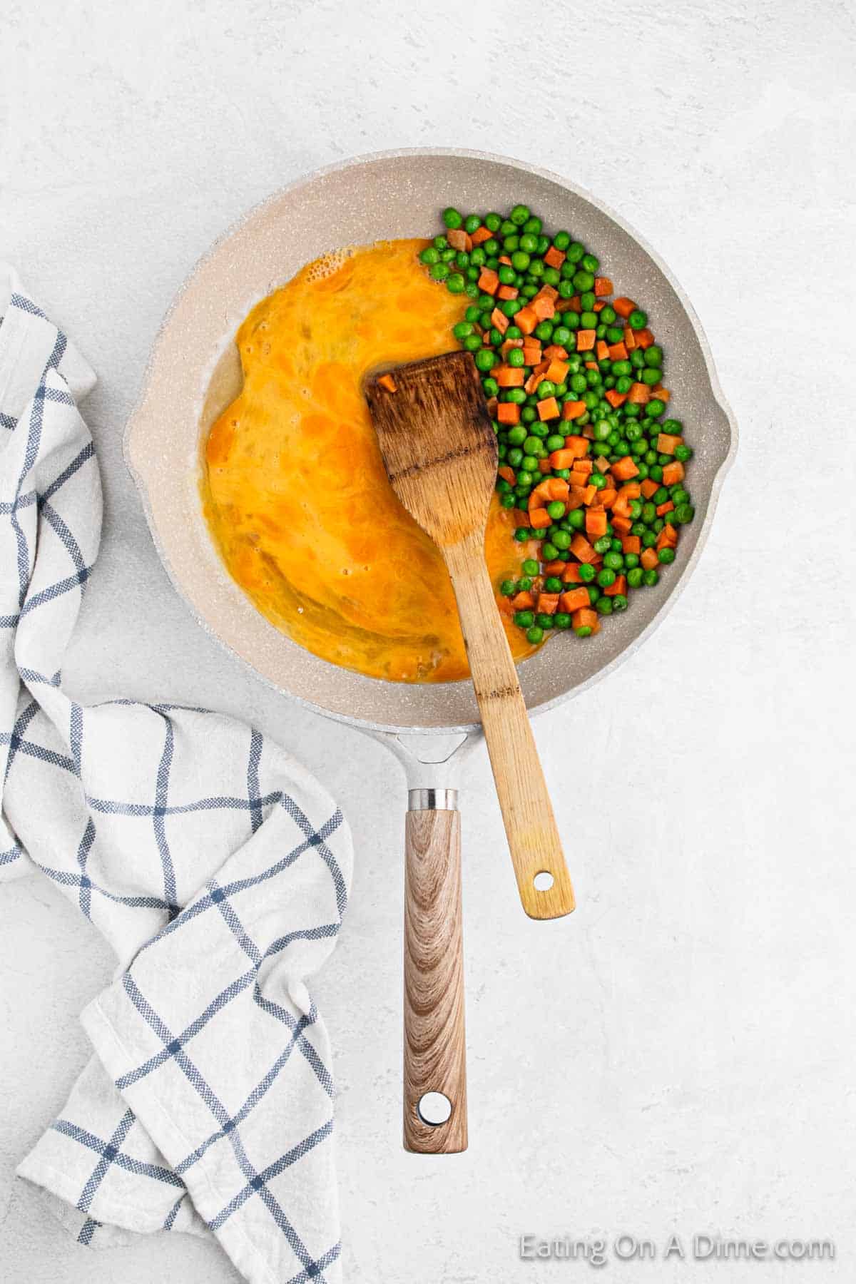 A frying pan sizzles with cauliflower fried rice, featuring eggs, peas, and carrots. A wooden spatula rests in the mixture, while a white and blue checkered towel lies nearby on a light surface.