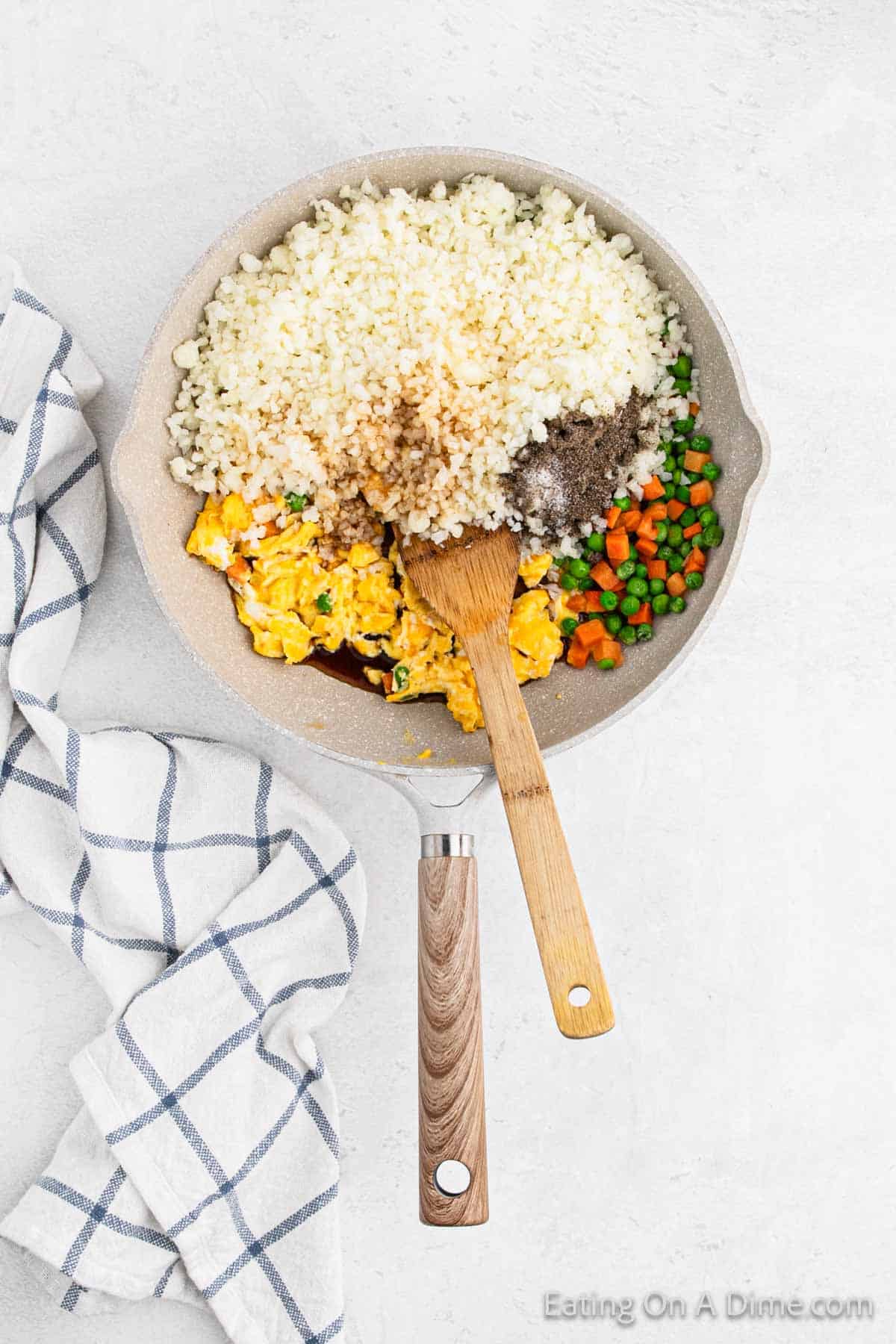 The skillet on a light surface holds a vibrant mix of cauliflower fried rice, scrambled eggs, peas, and carrots tossed with seasonings. A wooden spoon rests inside, ready to serve. Beside the skillet lies a checkered white and blue towel.