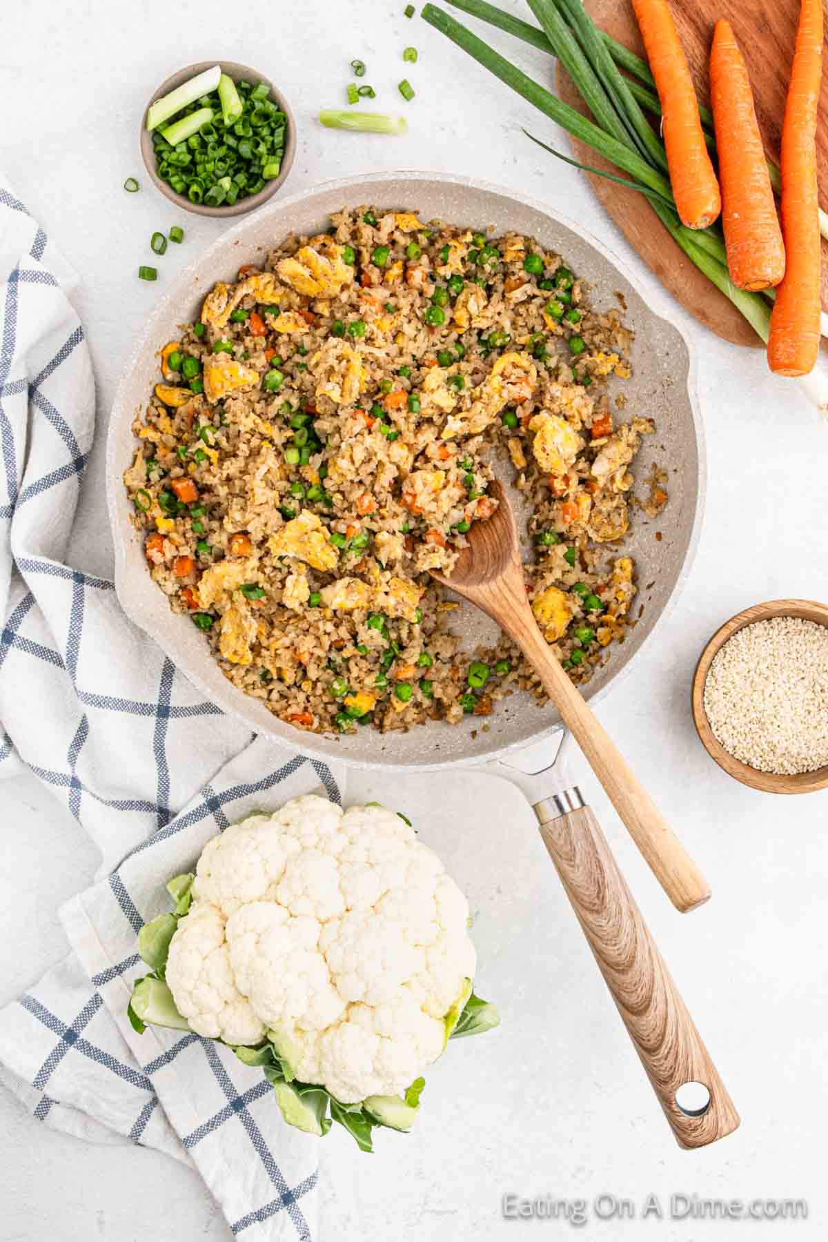 A bowl of cauliflower fried rice with peas, carrots, and scrambled eggs is garnished with chopped green onions. A wooden spoon rests in the bowl. The surroundings include raw carrots, a whole cauliflower, a small bowl of green onions, and sesame seeds.