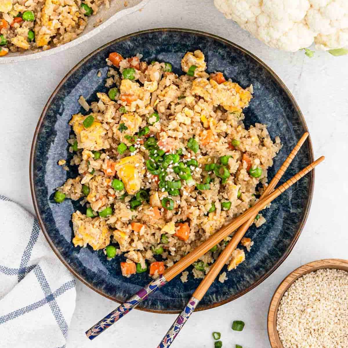 A plate of cauliflower fried rice with chopped vegetables and scrambled eggs, garnished with sesame seeds and green onions. Blue patterned chopsticks rest on the plate beside a small bowl of sesame seeds. A white napkin is partially visible, enhancing the vibrant dish's presentation.