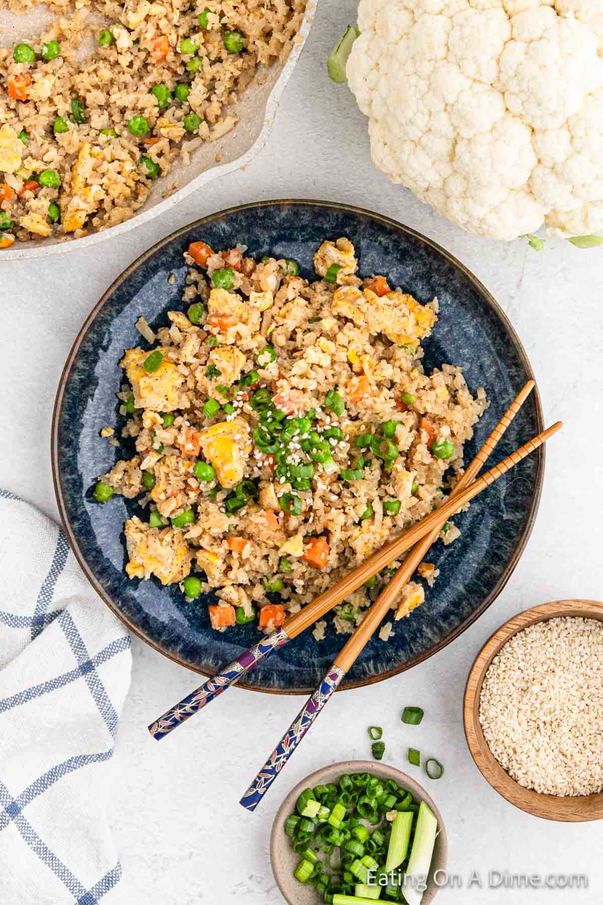 A plate of cauliflower fried rice with peas, carrots, and scrambled eggs is garnished with sesame seeds and green onions. Chopsticks rest on the side for an authentic touch. Nearby beckons a bowl of sesame seeds, sliced green onions, and a fresh head of cauliflower.