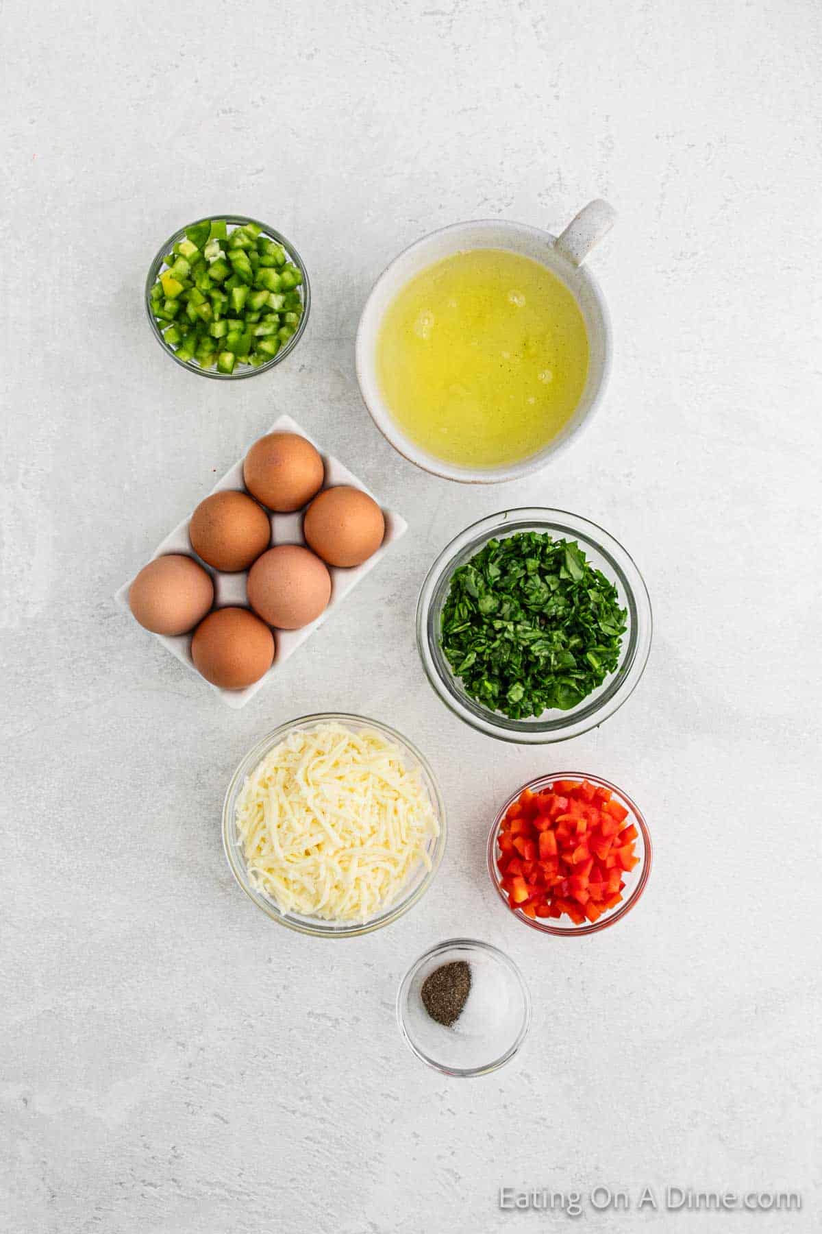 A top view of ingredients for an omelette or egg muffin cups recipe, featuring chopped green and red bell peppers, spinach, a bowl of egg whites, whole eggs, shredded cheese, and small bowls of salt and pepper, all arranged on a light gray surface.