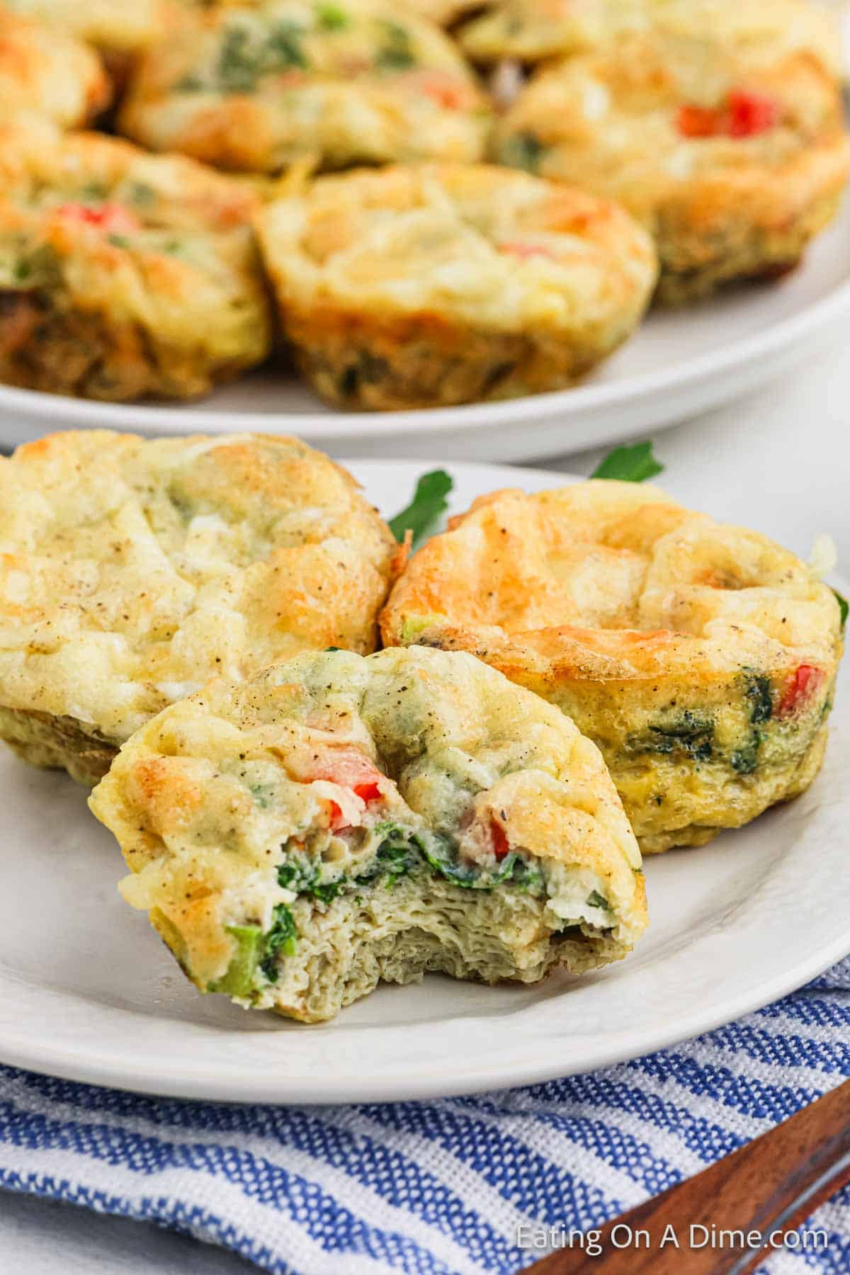 A plate of egg muffin cups with visible bits of vegetables like spinach, red pepper, and onions. One muffin has a bite taken out, revealing the savory recipe inside. The muffins are placed on a white plate over a blue and white striped cloth.