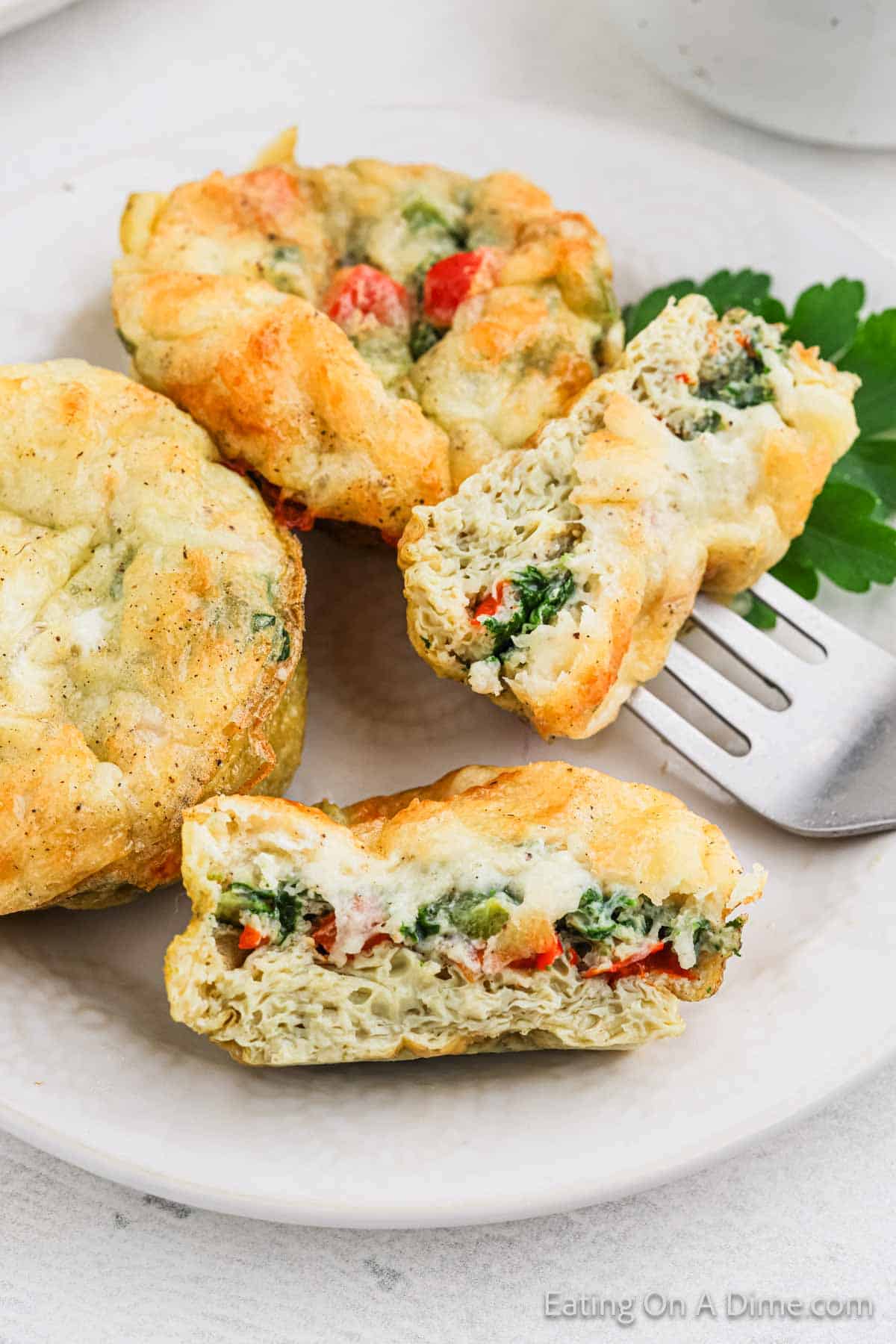 Three egg muffin cups on a white plate, filled with spinach and red bell peppers. Two muffins are whole, while one is cut in half to reveal the vibrant ingredients inside. A sprig of parsley is placed next to them, accompanied by a fork—an easy and colorful egg muffin cups recipe.