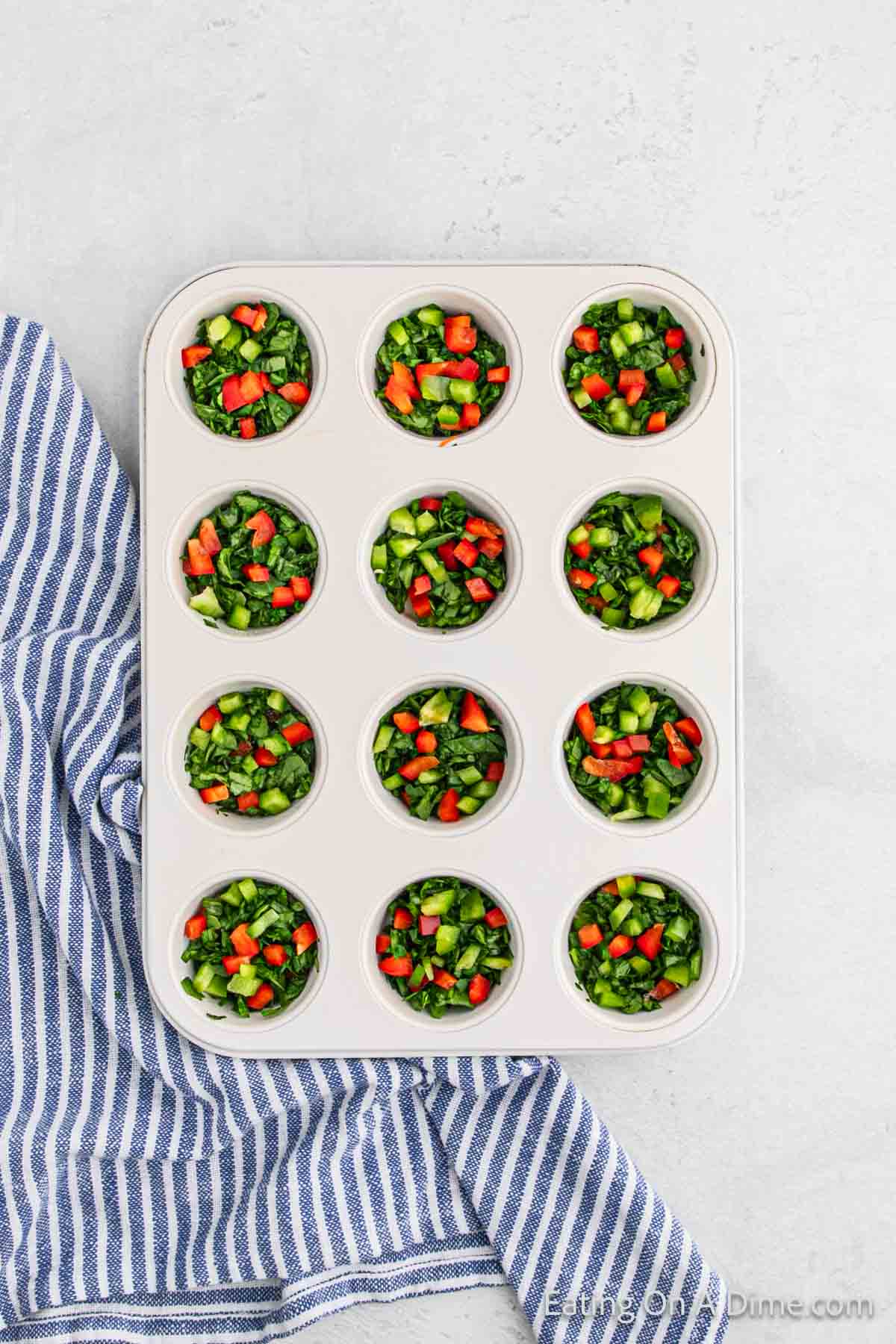 A muffin tin filled with diced vegetables, featuring green, red, and orange pieces, showcases an effortless egg muffin cups recipe on a light surface. A blue and white striped cloth partially covers the left side of the tin.