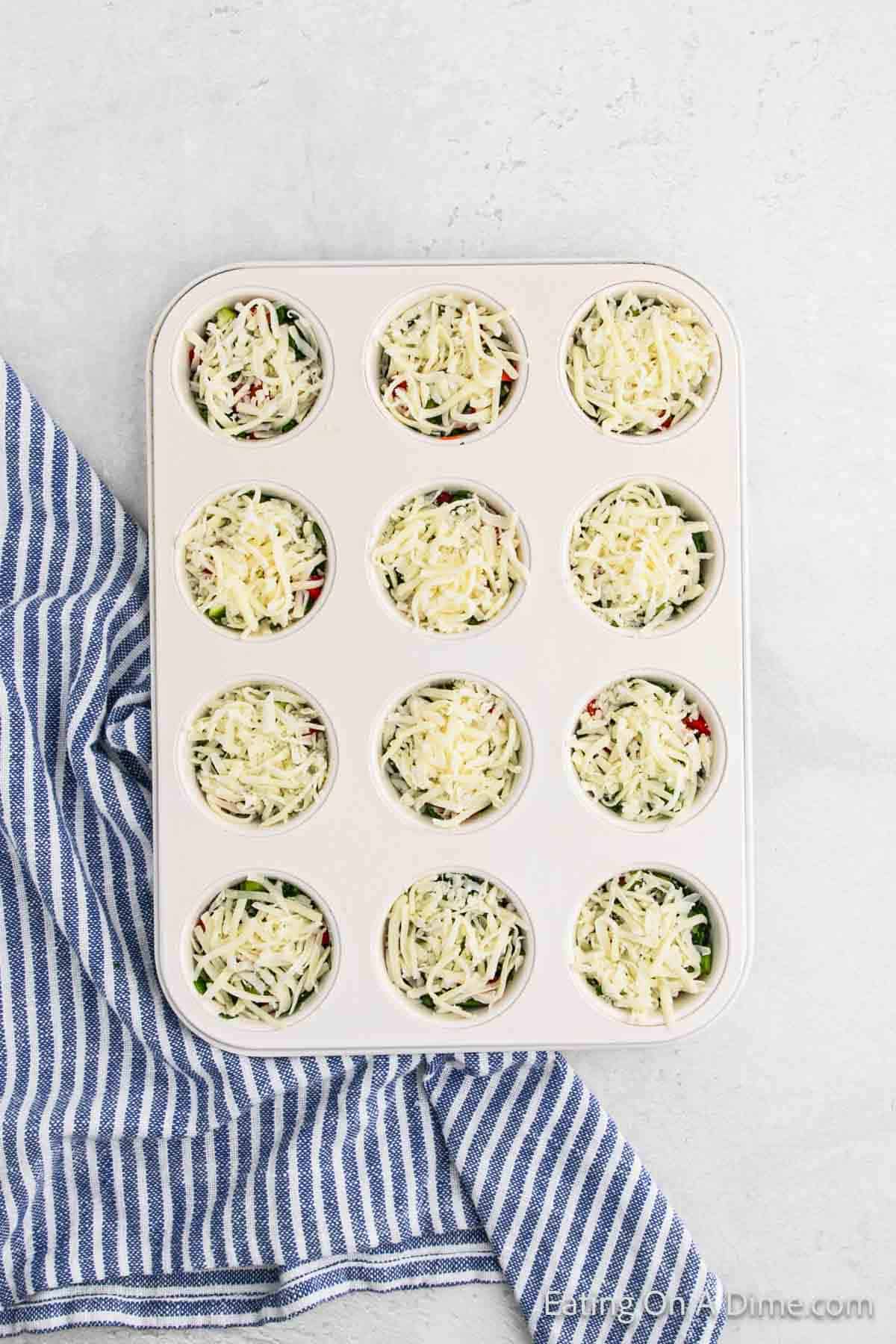 A muffin tray filled with uncooked egg muffin cups topped with shredded cheese rests on a blue and white striped cloth. The enticing muffins reveal various fillings peeking through the melted cheese, perfect for any recipe adventure.