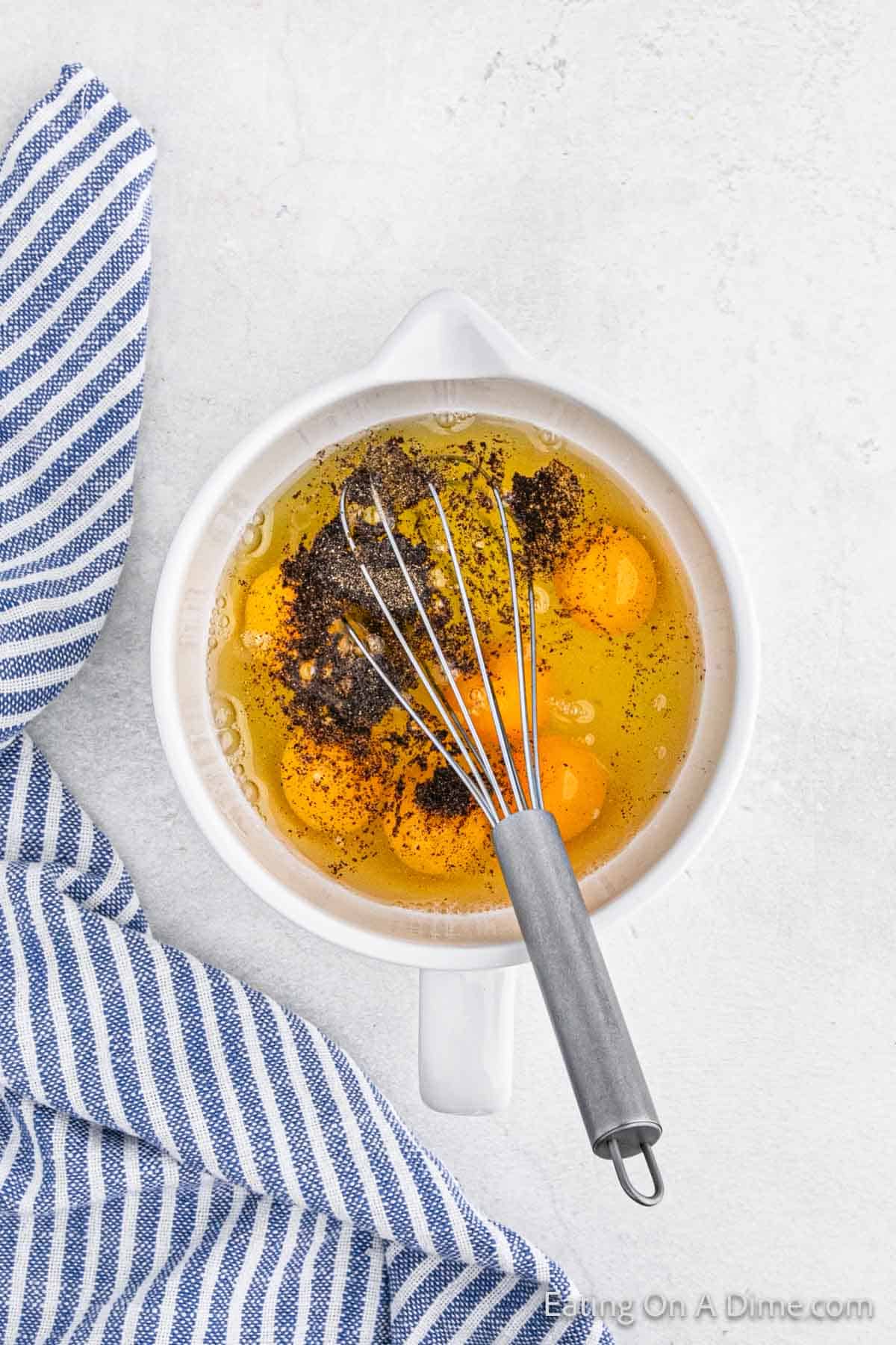 A white bowl containing cracked eggs and ground pepper is placed on a light surface, ready for an egg muffin cups recipe. A whisk rests on the bowl, while a blue and white striped cloth is draped beside, setting the stage for a delightful culinary creation.