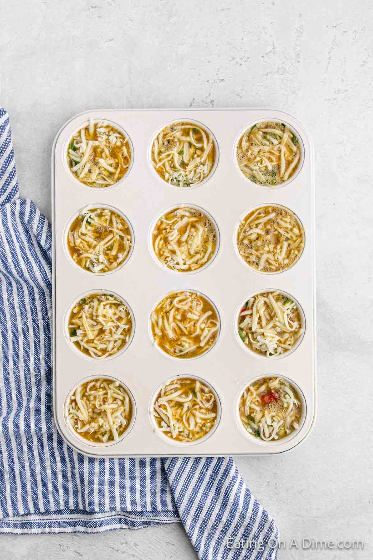 A muffin tin filled with an egg muffin cups recipe is on a light surface. Each cup contains a mixture of eggs, shredded cheese, and vegetables. A blue and white striped cloth is partially visible on the left.