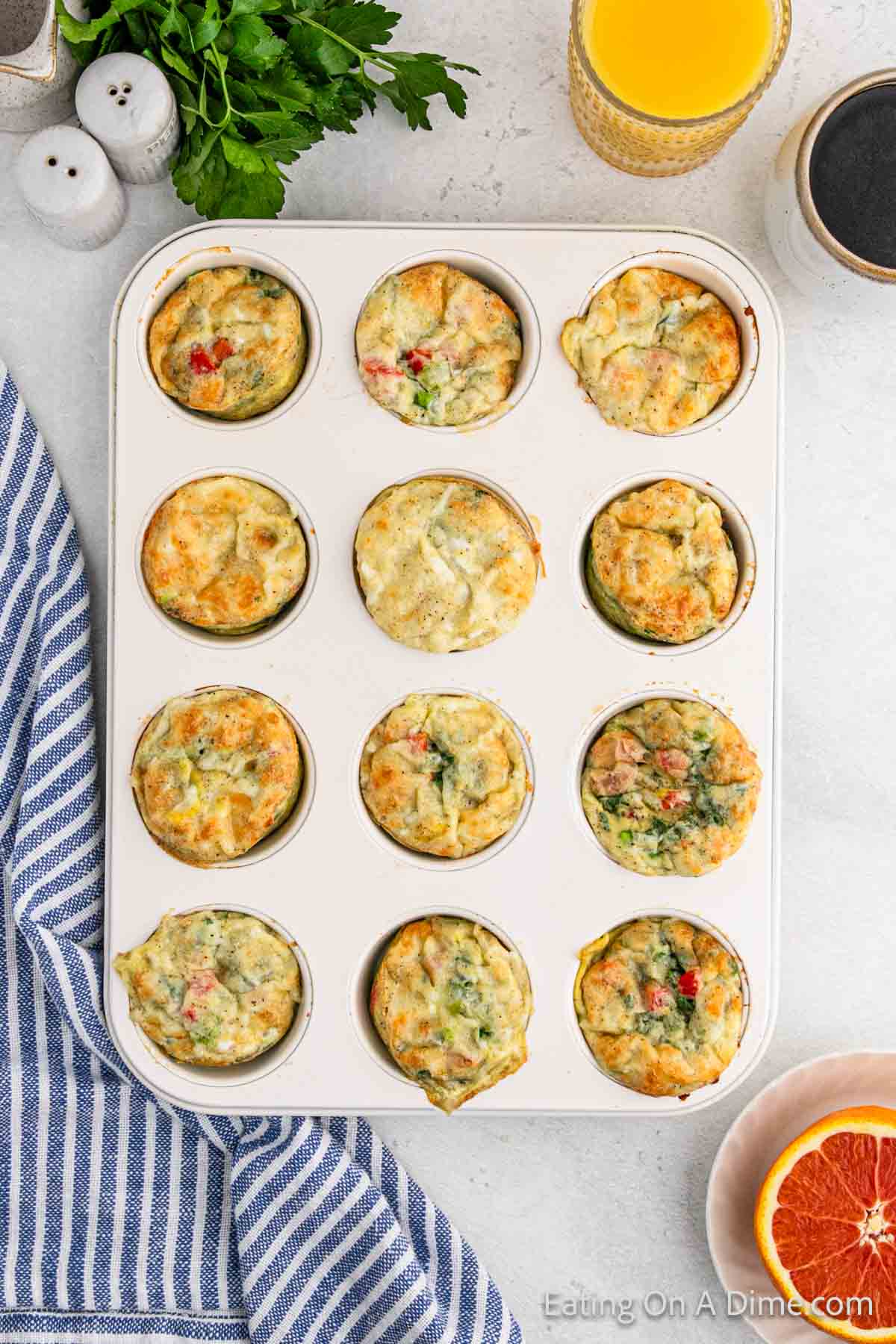 A muffin tray filled with assorted egg muffin cups, featuring visible vegetables. Nearby are a striped napkin, a glass of orange juice, parsley, salt and pepper shakers, and half a grapefruit on a light-colored countertop—perfect for trying out your favorite egg muffin cups recipe.